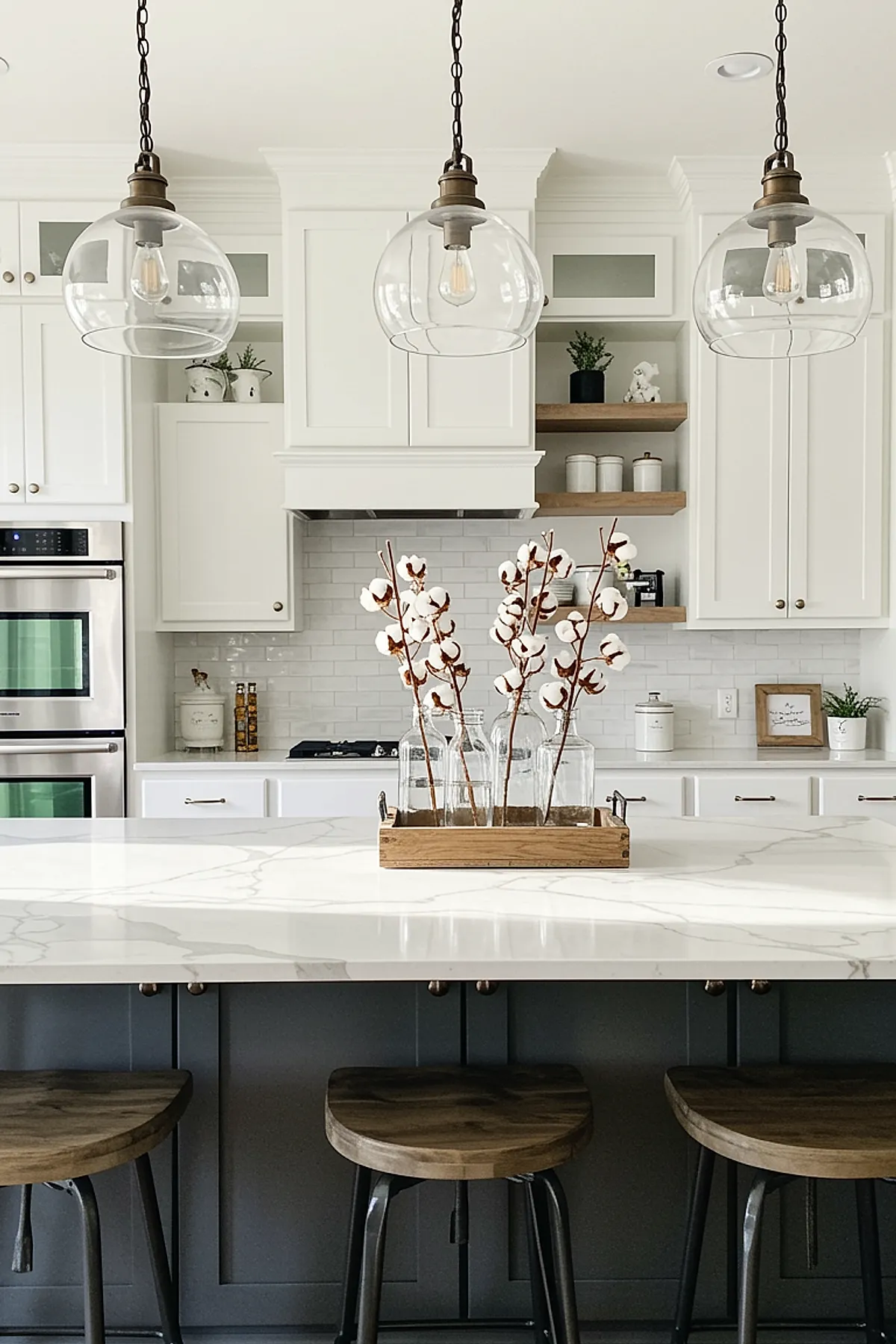Clear glass pendant lights over a white kitchen island with wooden stools and cotton plant decor.