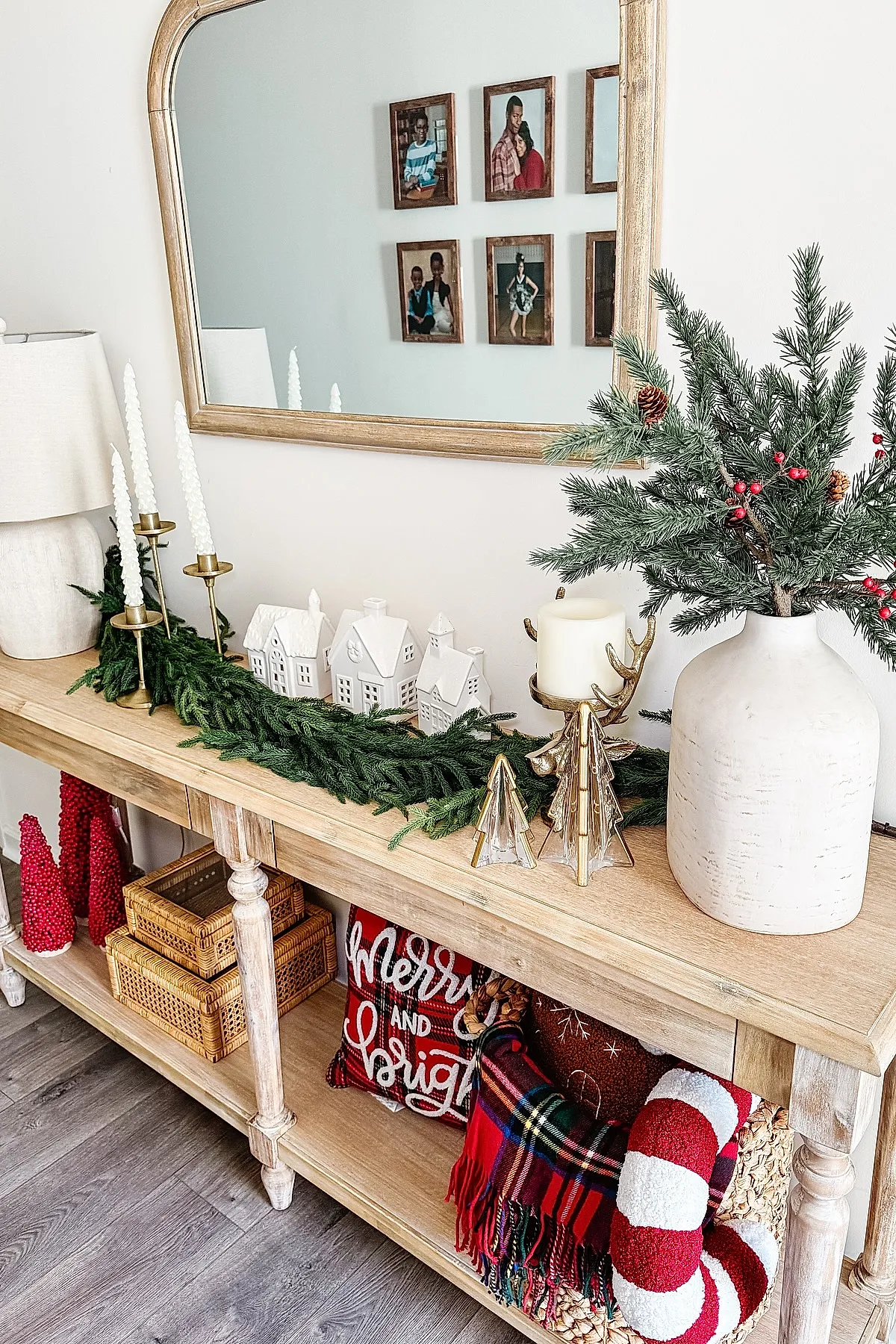 Light wooden console table with green garland, white twisted candles in gold holders, ceramic houses, pine branches with red berries in a white vase, wicker baskets, plaid throw, and festive pillows on wood floor.