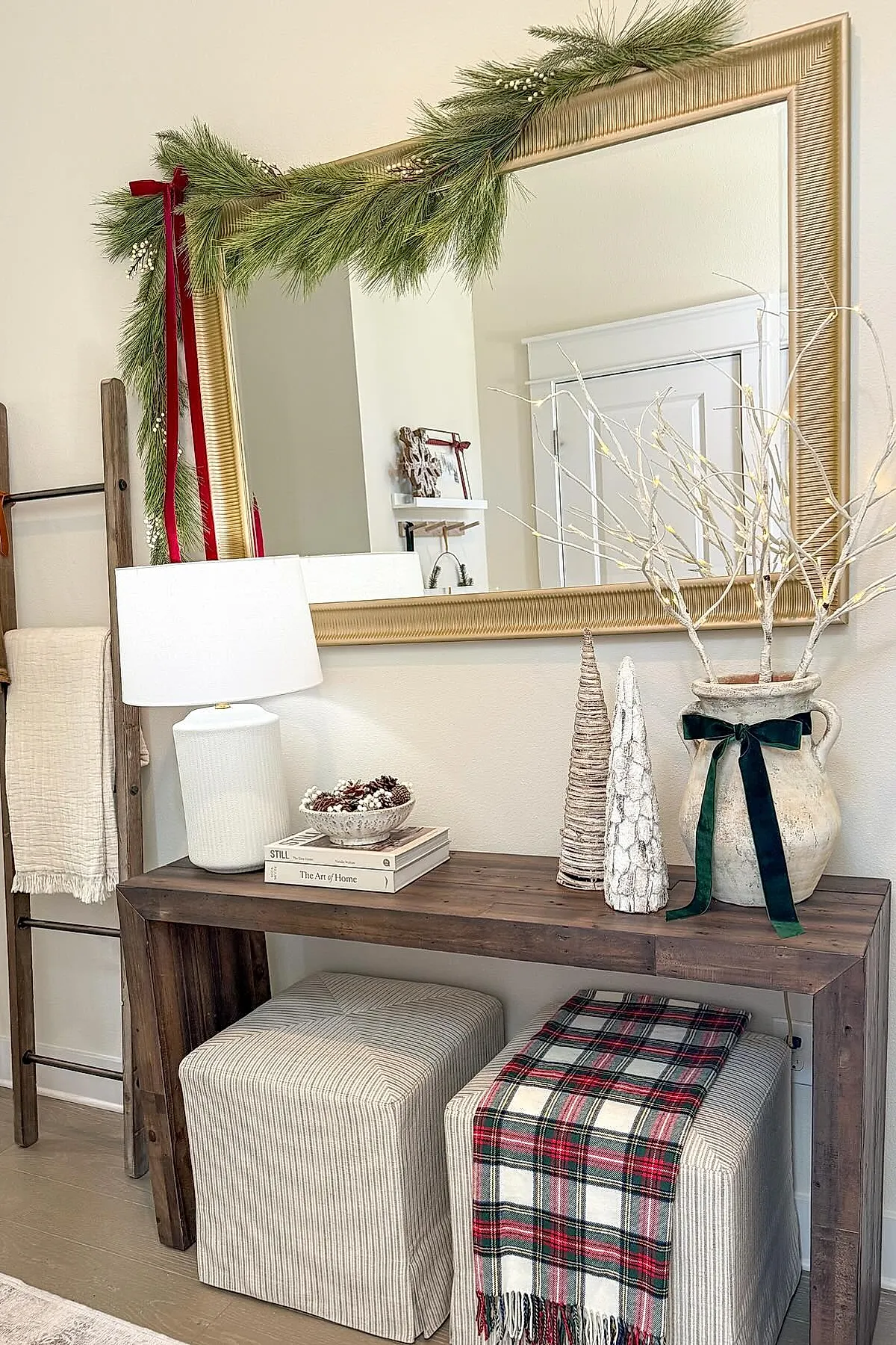 Console table with gold-framed mirror decorated with pine garland and red velvet ribbons, white lamp, pinecone bowl, cone-shaped trees, ceramic vase with white branches tied with green velvet ribbon, and striped ottomans underneath.