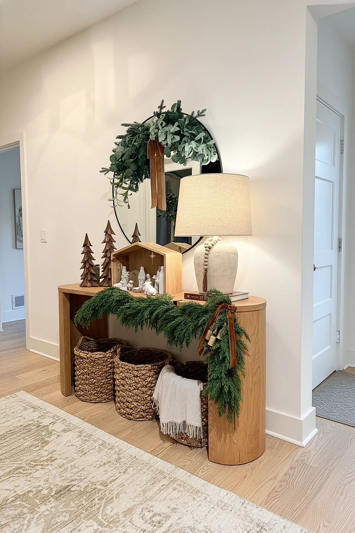 Light wooden console table with natural green garland tied with brown velvet ribbons and bells, beige lamp, wooden nativity scene, carved wooden Christmas trees, stacked books, and woven baskets below.
