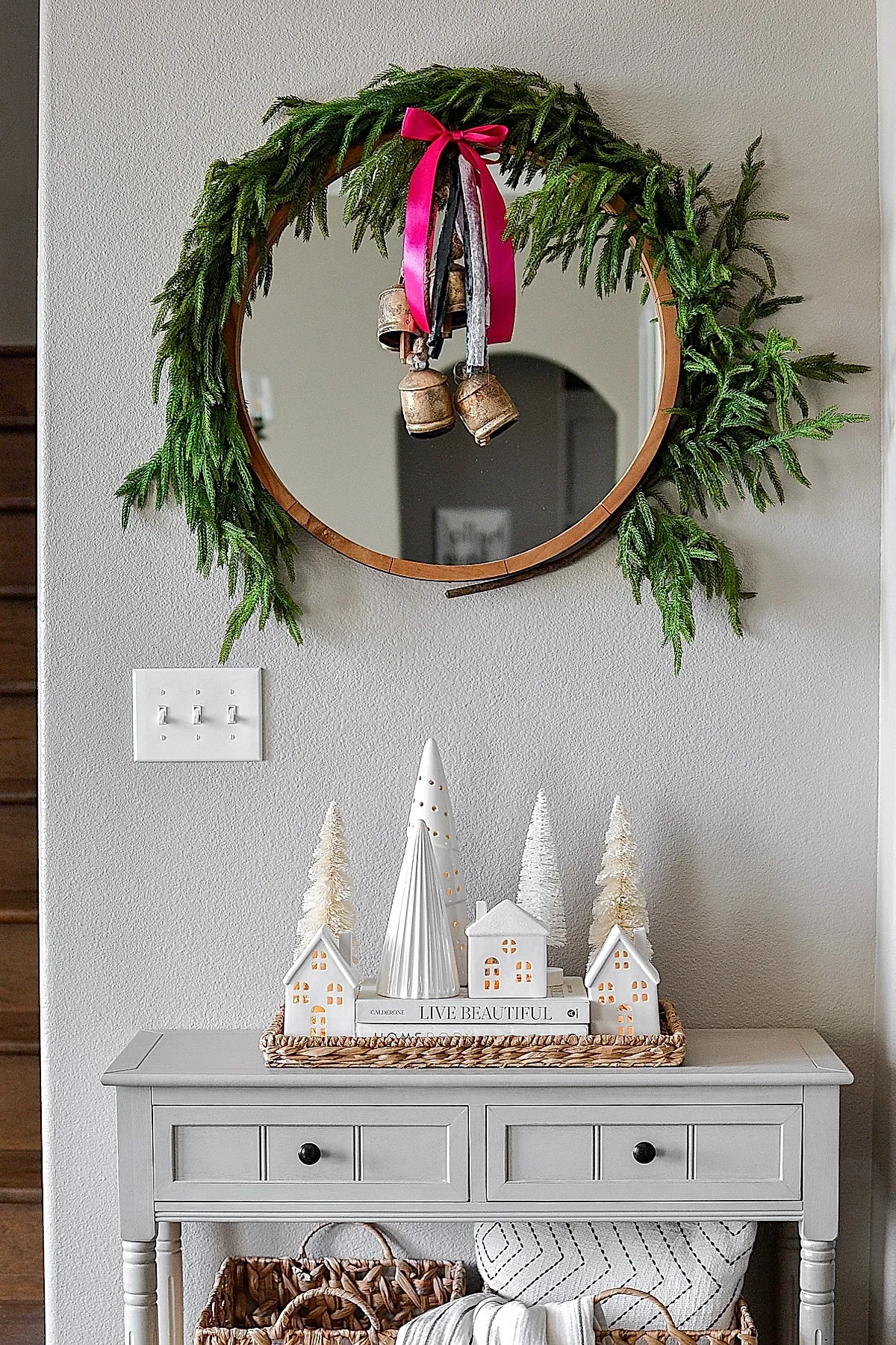 Gray console table with woven tray holding white ceramic houses and bottle brush trees on stacked books, round wooden-framed mirror with pine garland and vintage jingle bells tied with pink ribbon.