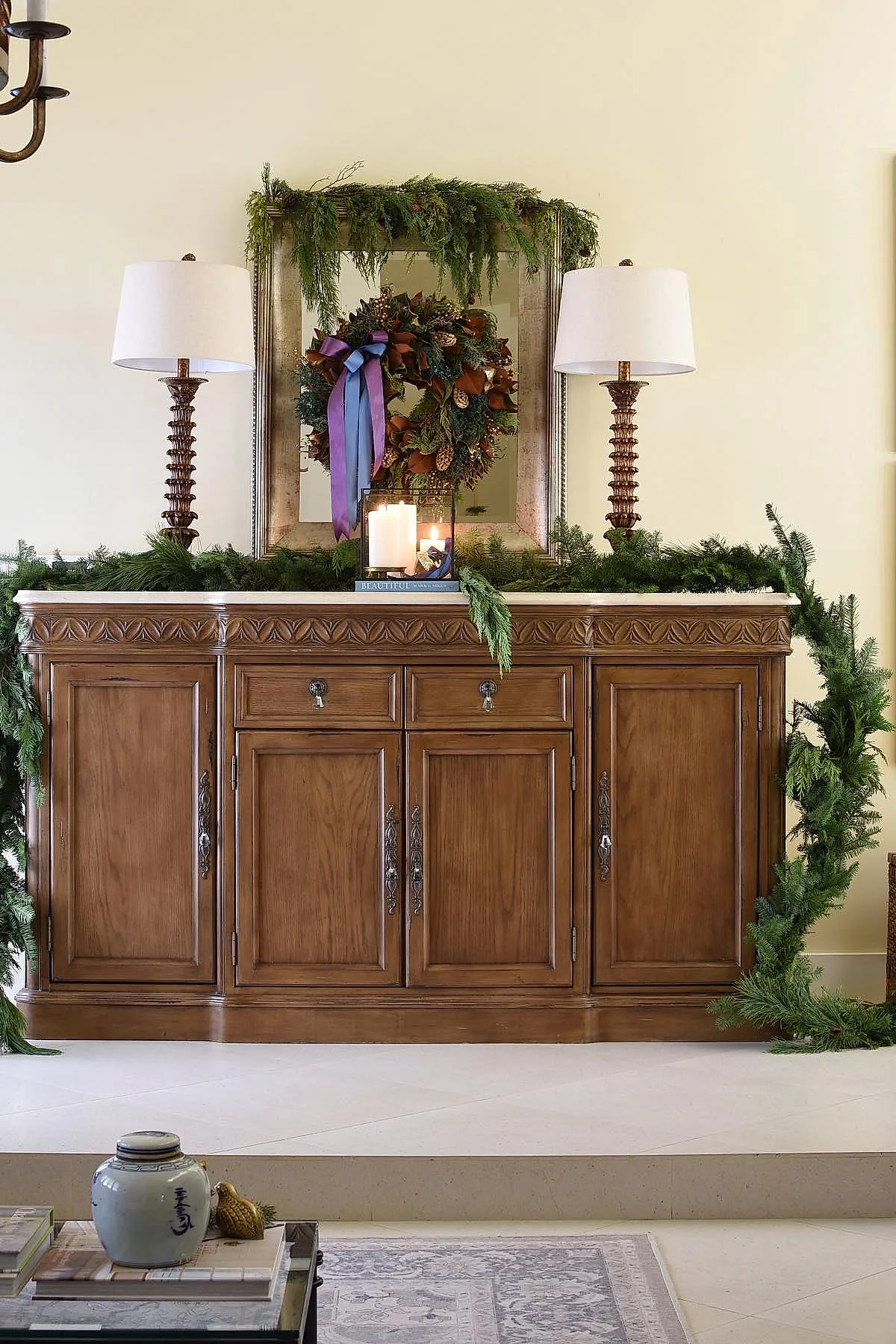 Wooden console table with lush green garland, two tall lamps with white shades, large framed mirror with pine wreath featuring brown leaves and pinecones tied with purple and blue ribbons, white pillar candles in glass lantern on books.