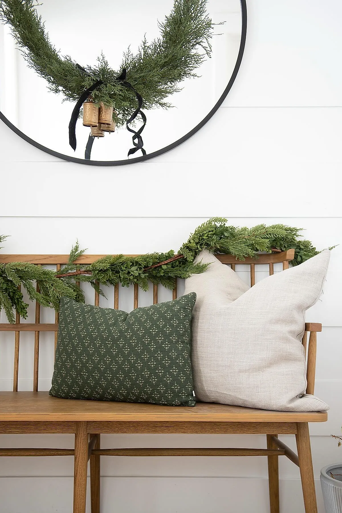 Wooden entryway bench with green garland draped over backrest, dark green patterned pillow, large beige pillow, white shiplap wall, round mirror with wreath accented by black ribbon and small bells.