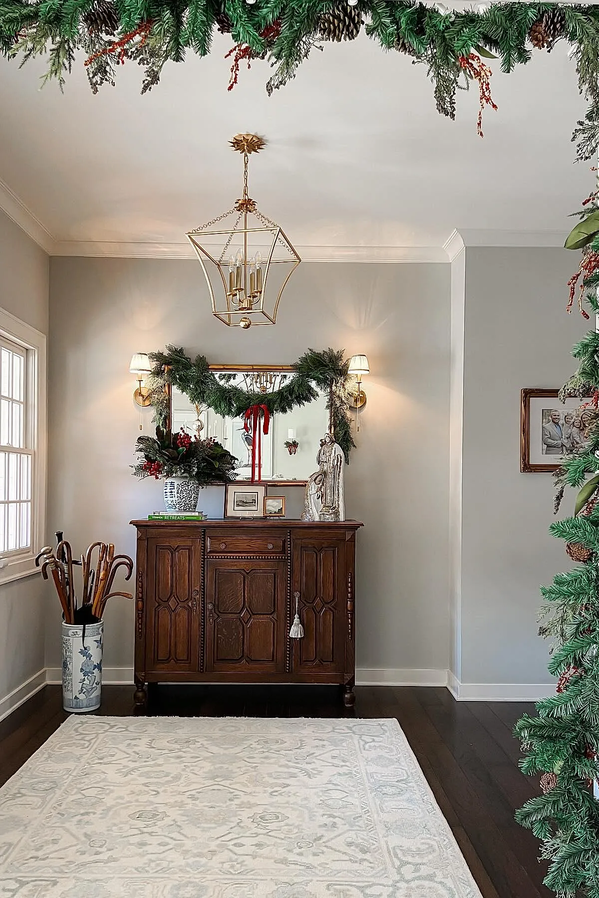 Vintage wooden console table with lush greenery garland over large gold-framed mirror, red berries, white vase with floral arrangement, framed photos, silver nativity figurine, gold wall sconces, umbrella stand with wooden handles on dark hardwood floors.