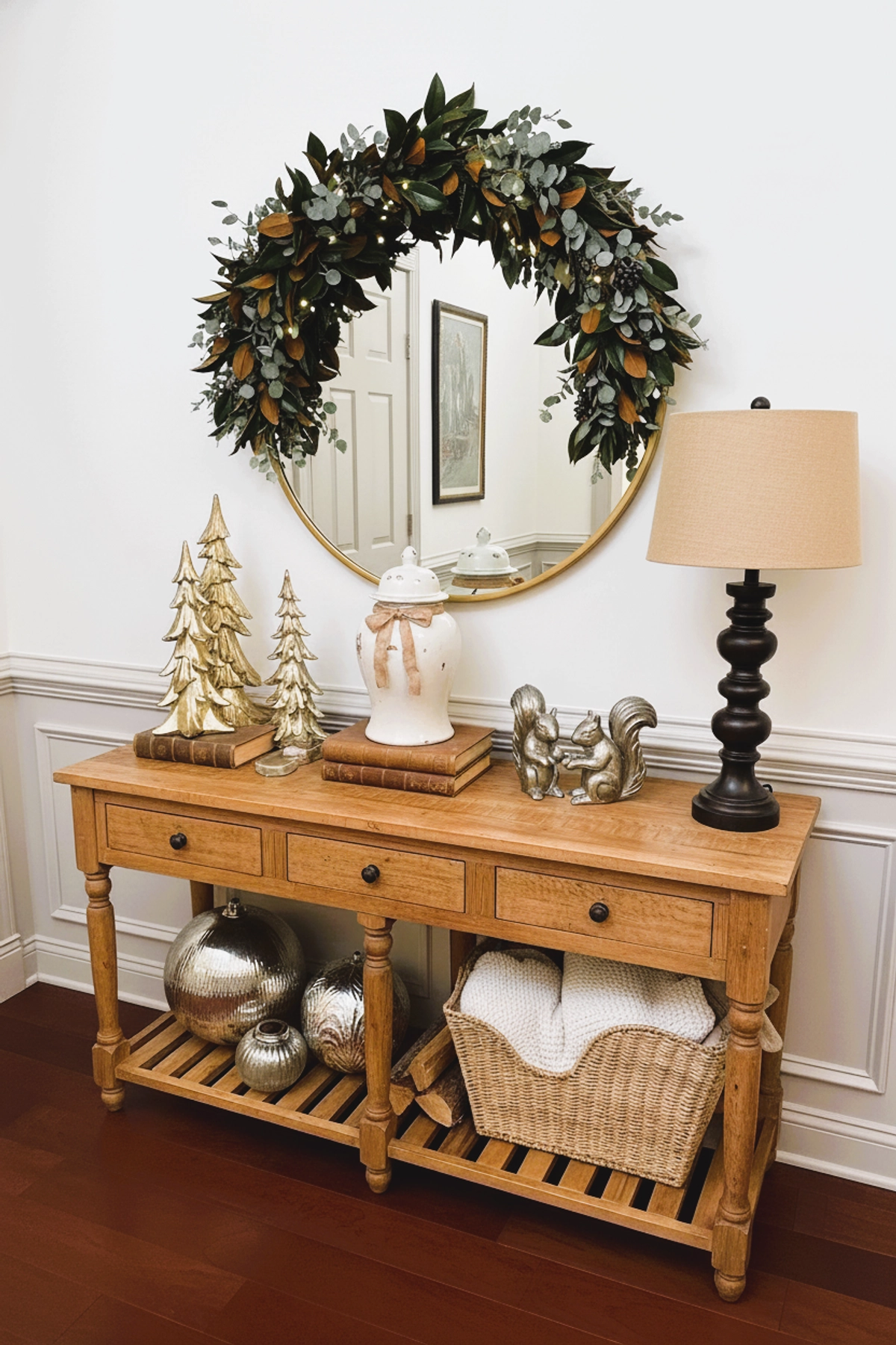 Christmas entryway with magnolia and eucalyptus wreath over round mirror, wooden console table with gold Christmas trees, stacked books, ceramic jar with ribbon, metallic ornaments, wicker basket with white blankets.