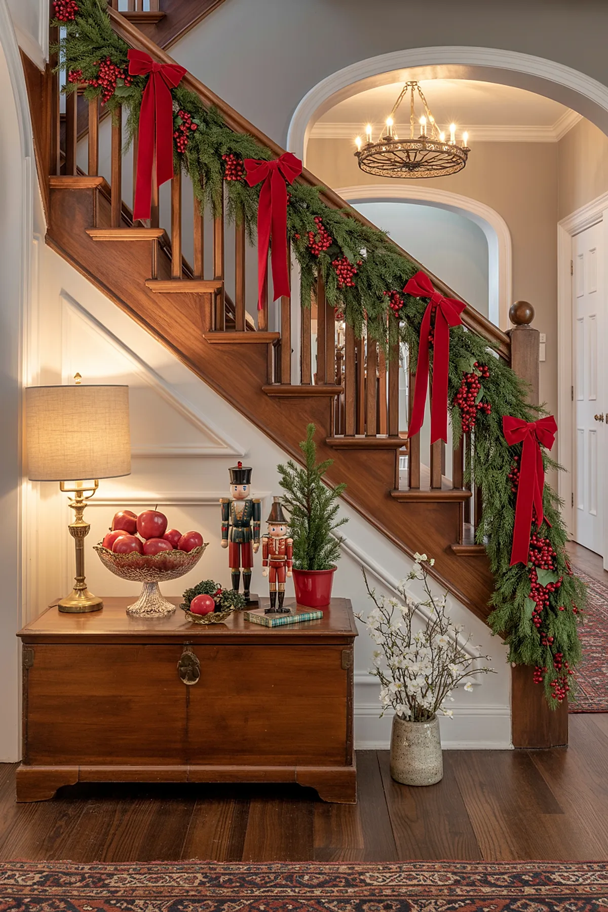 Wooden staircase decorated with green garland, red berries, large red velvet bows; wooden chest with lamp, bowl of red apples, nutcracker figurines, small potted evergreen, vase with white blossoms, beige walls, dark hardwood floors.