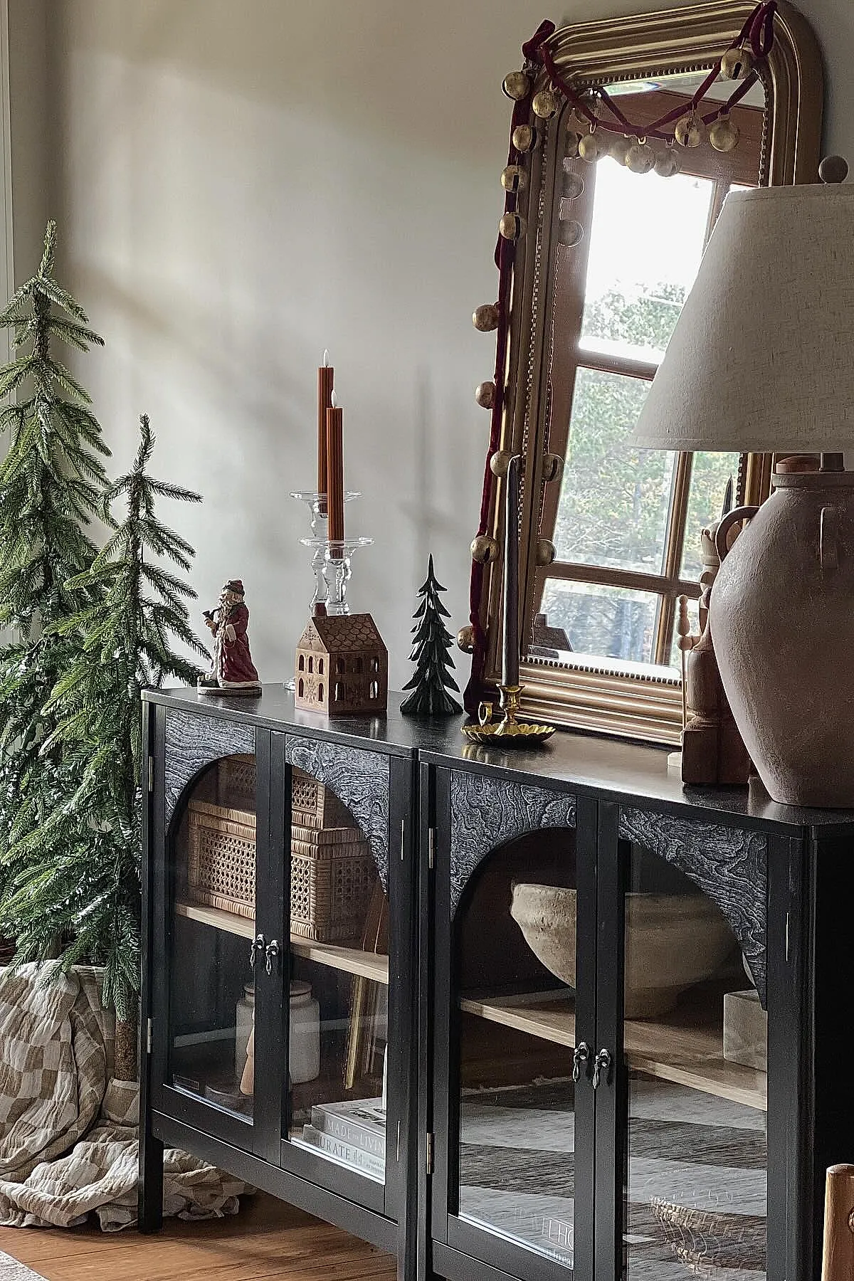 Black console table with glass doors holding wicker baskets and books, topped with tall brown candles in clear holders, small green Christmas trees, ceramic Santa figurine, large gold-framed mirror with red ribbon garland, beige lamp, green Christmas tree wrapped in neutral cloth.