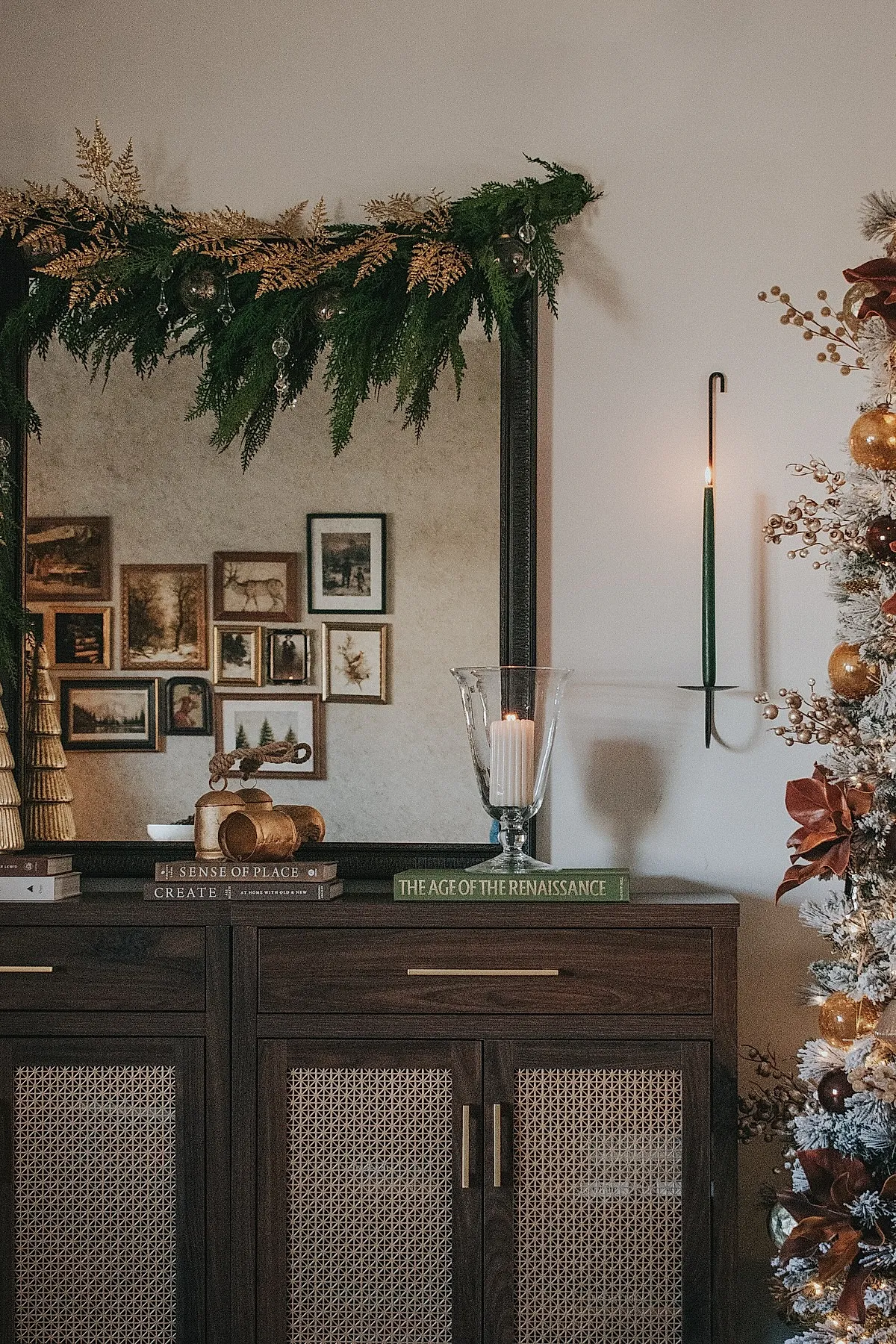 Dark wooden console table with green garland accented by gold leaves and glass ornaments, large candle in glass hurricane holder on green book, mirror reflecting vintage artwork, decorated white Christmas tree with brown and gold ornaments.