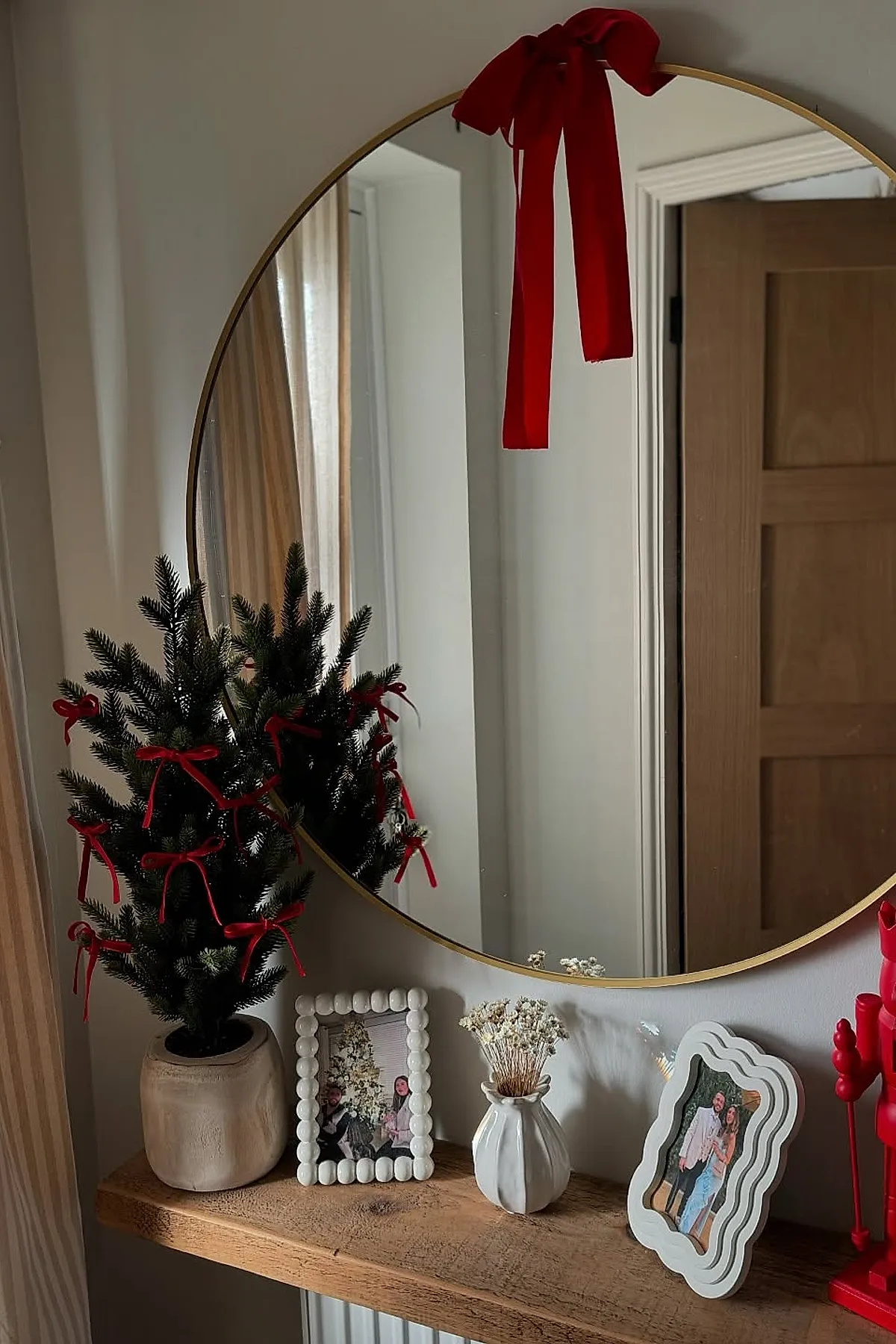 Wooden entryway shelf with small pine tree decorated with red velvet bows in beige ceramic pot, large round mirror with red bow, white photo frames, small white vase with dried flowers, tall red decorative candle.