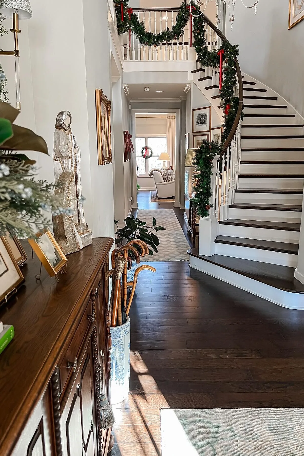 Curved white staircase with dark wood steps decorated with green garlands and red ribbons, wooden console table with framed pictures and decorative statue, umbrella stand with walking sticks, hallway leading to bright living room with wreath on window.