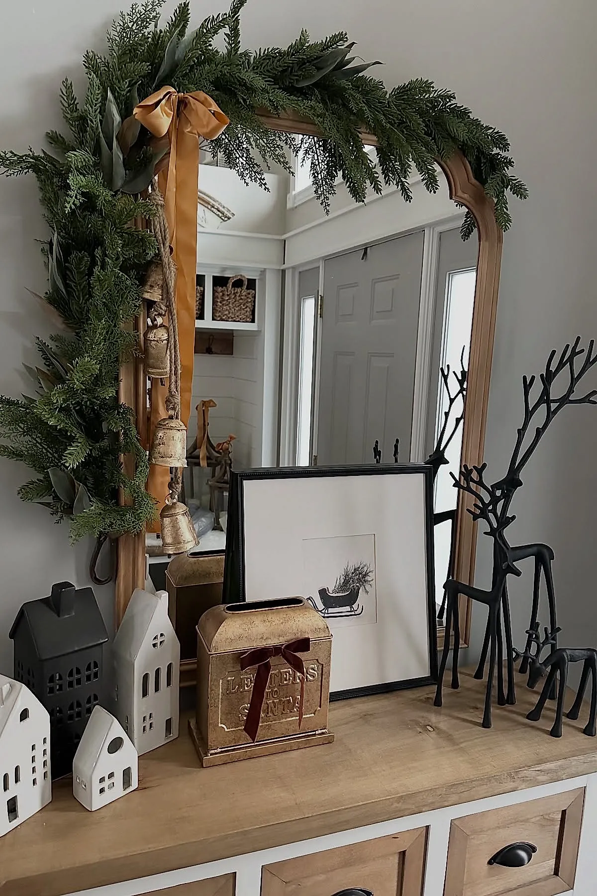 Wooden console table with vintage metal mailbox labeled 'Letters to Santa' tied with dark red ribbon, black metal reindeer figurines, ceramic house lanterns, framed sleigh print, large mirror with green garland and golden bells tied with orange satin ribbon.