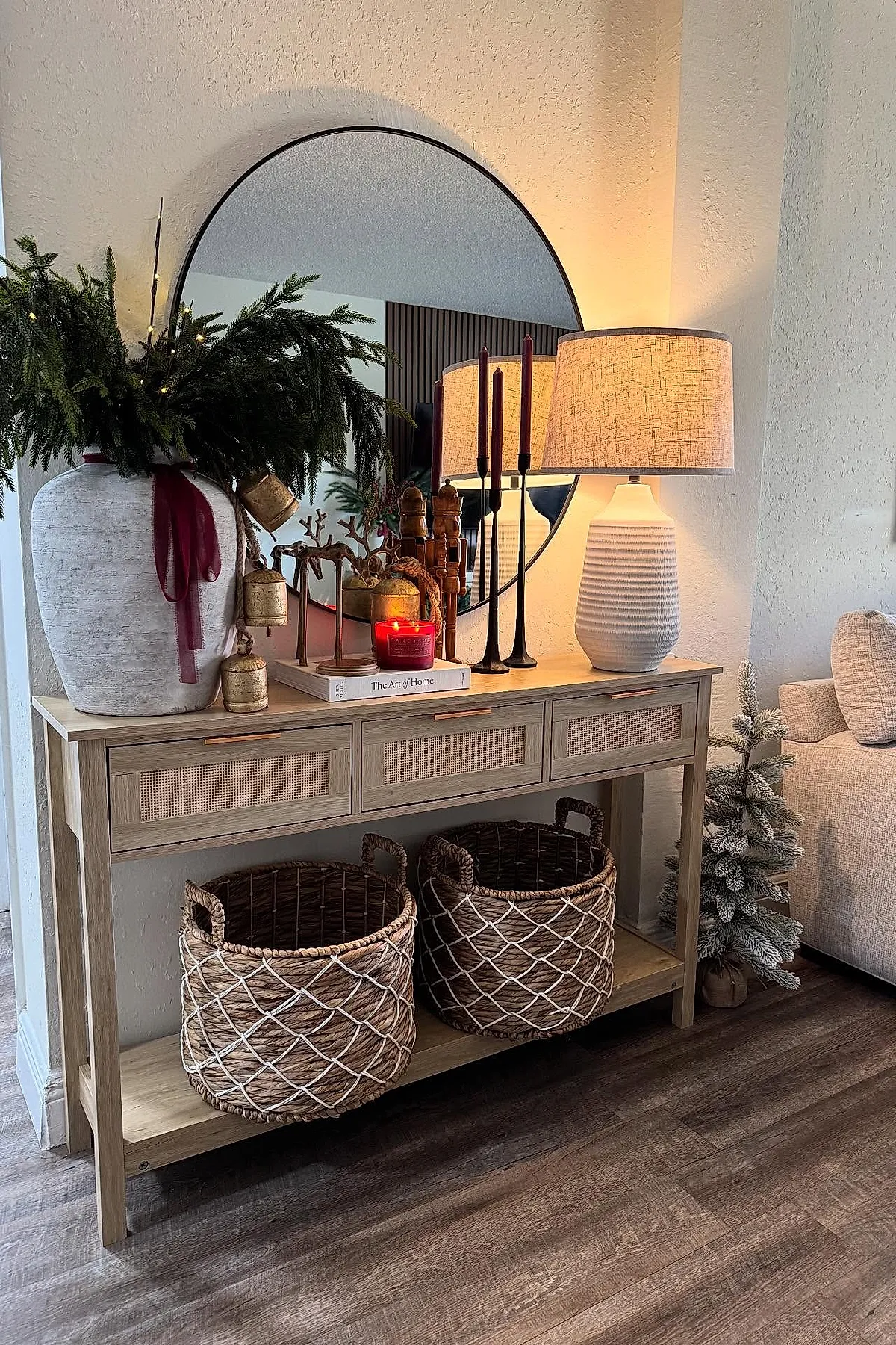 Light wooden console table with large vase of pine branches and gold bells, tall dark red candles, red scented candle on book, white textured lamp, woven baskets, small snowy Christmas tree beside beige sofa.