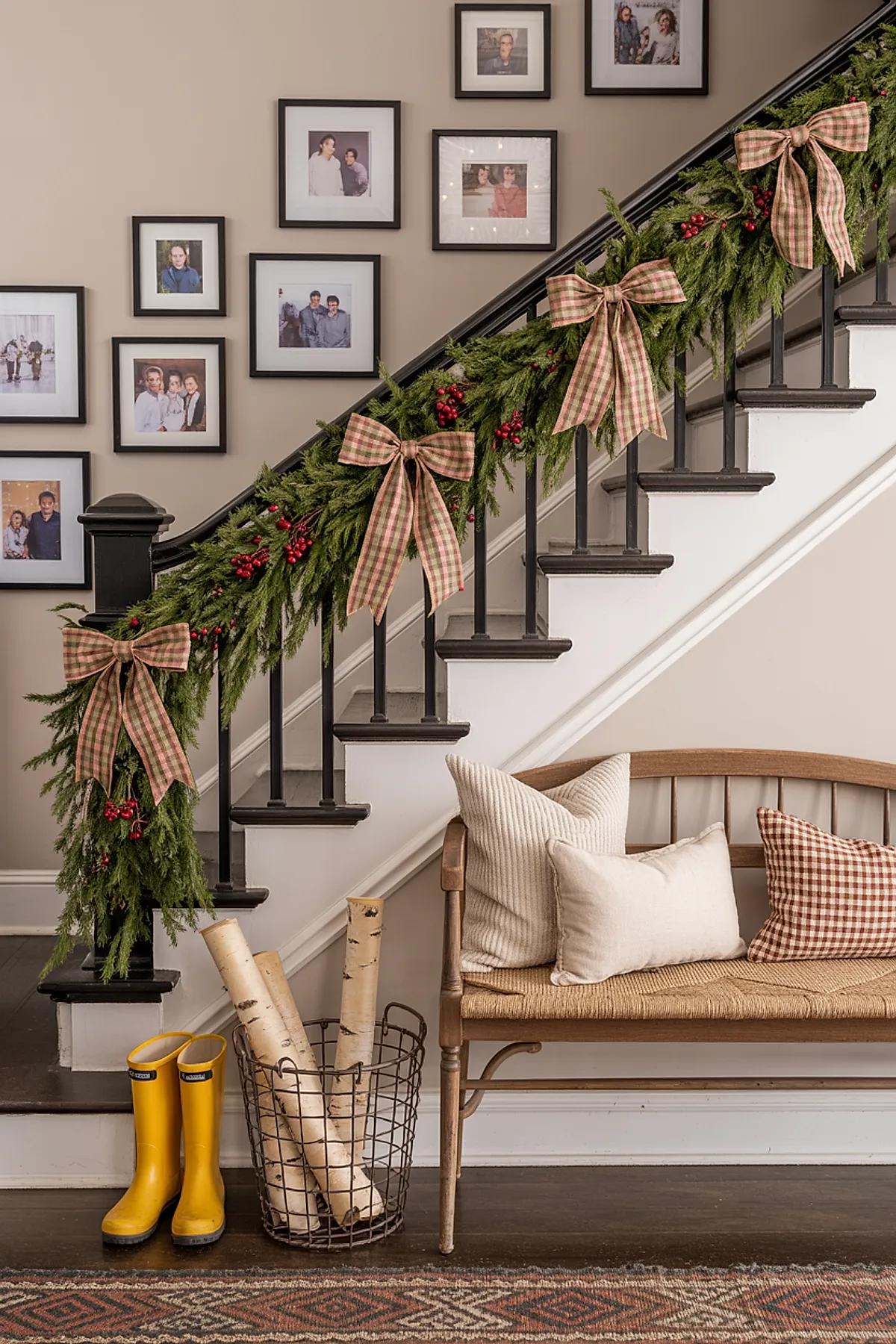 Black staircase banister decorated with green pine garland, red berries, large plaid bows in beige and red, wooden bench with woven seat holding cream and gingham pillows, wire basket with birch logs, yellow rain boots on dark hardwood floors.