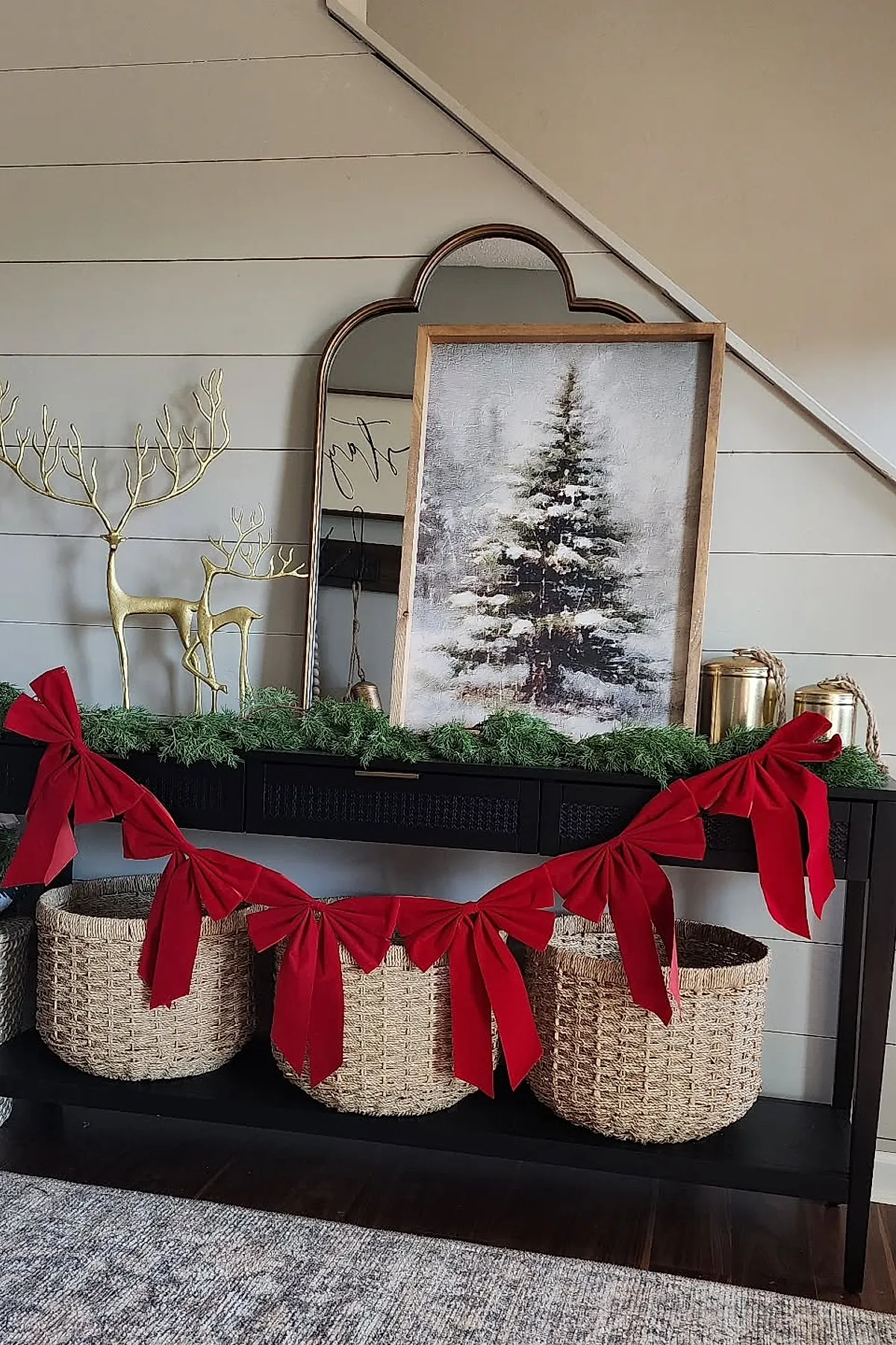 Black console table with green garland and large red velvet bows, gold metal reindeer figurines, framed winter tree painting, scalloped mirror, gold lanterns, woven baskets on dark hardwood floors.