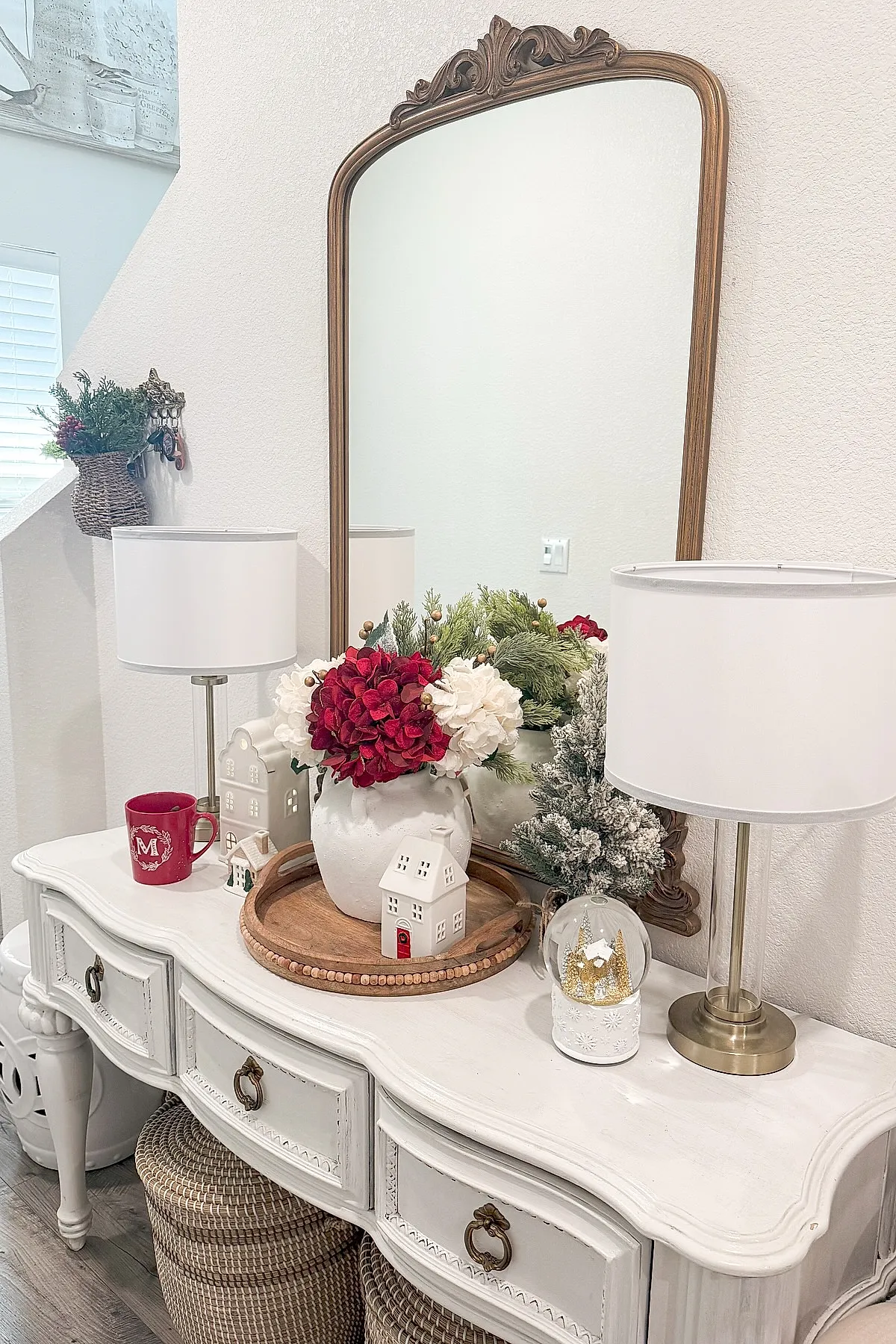White vintage-style console table with large white vase holding red and white flowers and greenery on wooden tray, ceramic house figurines, snow globe with golden trees, frosted pine tree, modern lamps with white shades, beige wall, ornate wooden framed mirror.