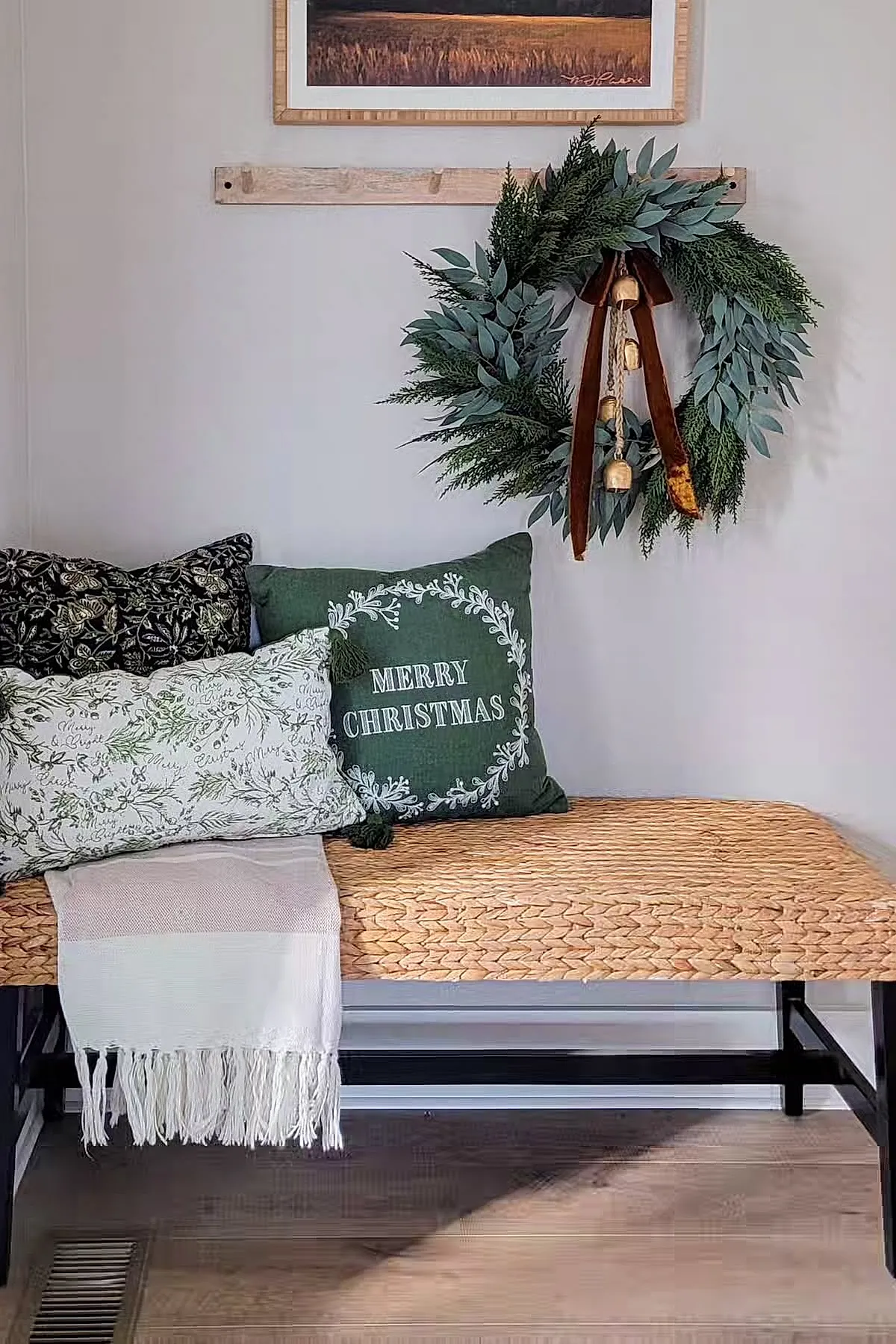 Woven natural fiber bench with three festive pillows: dark green 'Merry Christmas' pillow, white pillow with green botanical prints, black floral pillow; fresh green wreath with gold bells and rust-colored velvet ribbon hanging above.