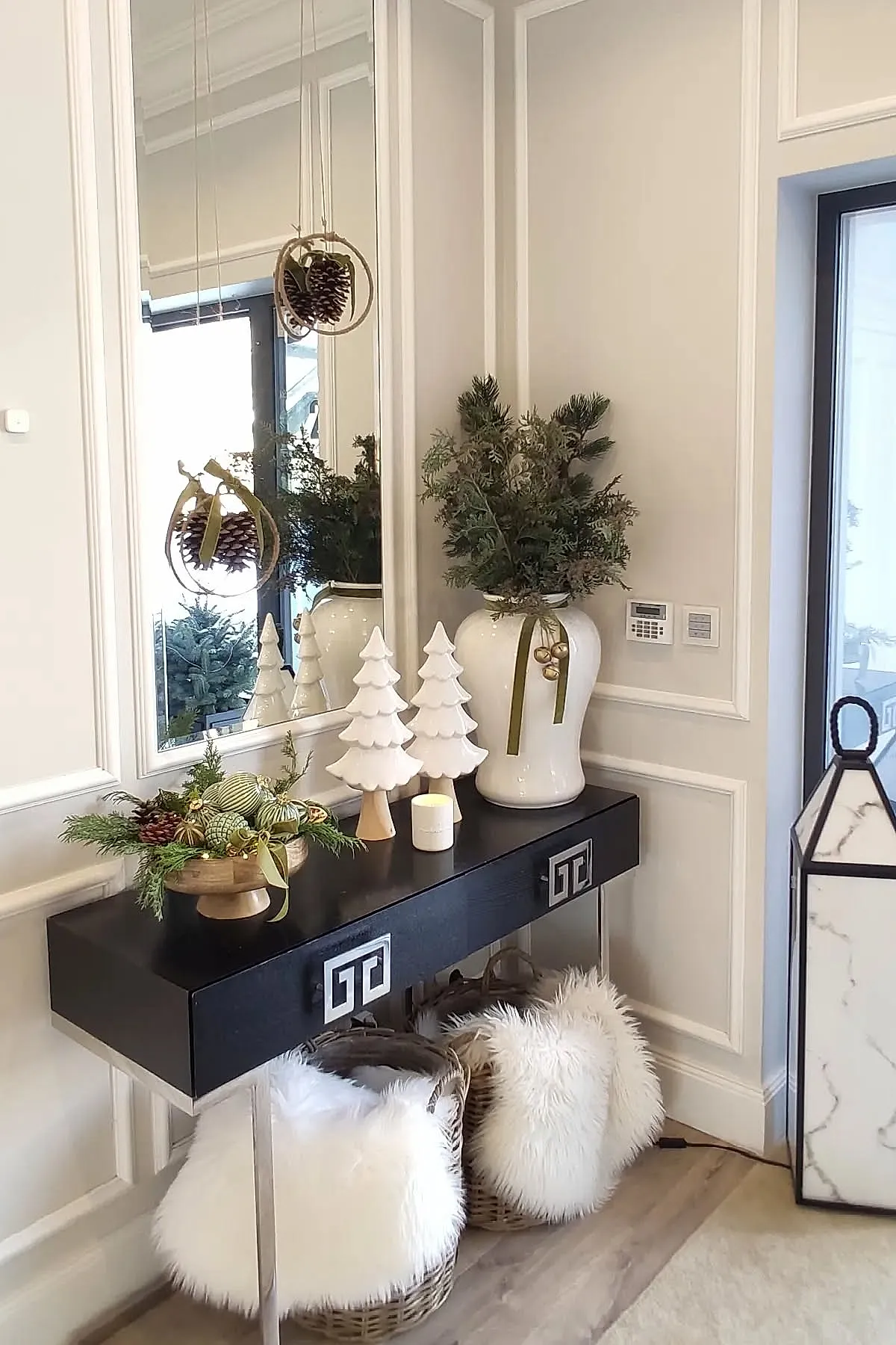 Modern black console table with white ceramic Christmas tree figurines, small candle, wooden bowl with green ornaments and pinecones, large white vase with pine branches tied with gold bells, tall wall mirror, light walls with decorative molding.