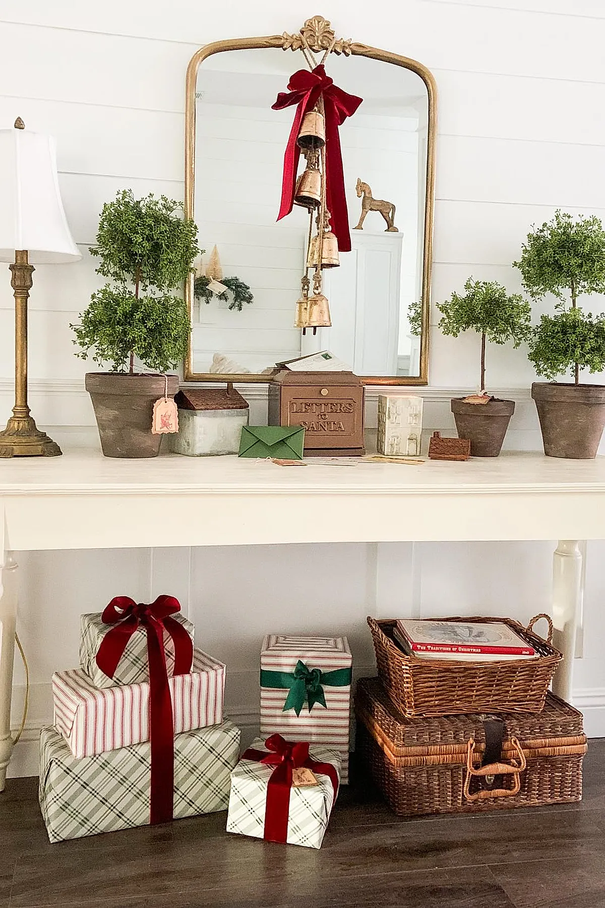 White console table with gold-framed mirror, small potted topiary trees, brown metal 'Letters to Santa' mailbox, ceramic house figurines, green envelope, wrapped presents in plaid and striped paper with red and green ribbons, woven baskets on dark wood floor.