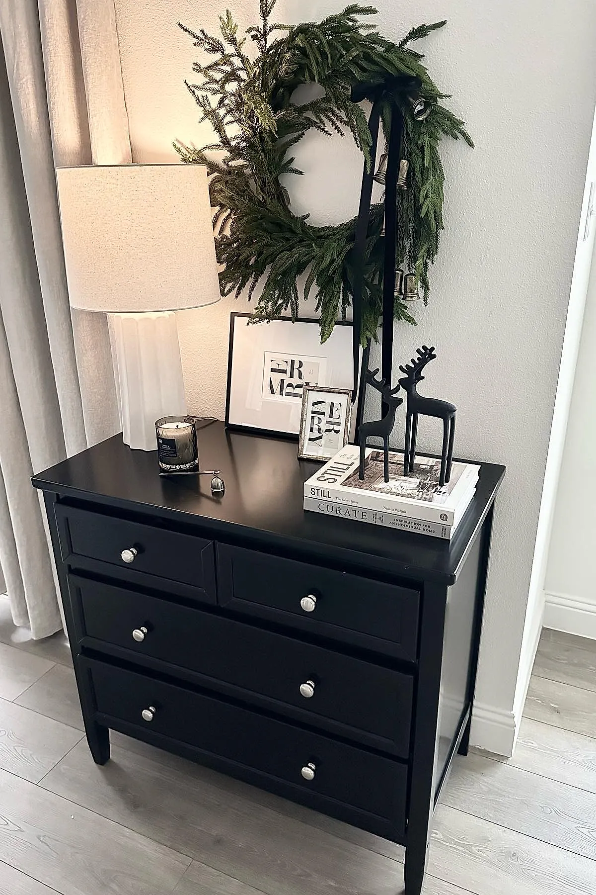 Black wooden console table with large green wreath adorned with silver bells and black ribbon, two black reindeer figurines on stacked books, framed artwork, white lamp with beige shade, small candle in glass holder.