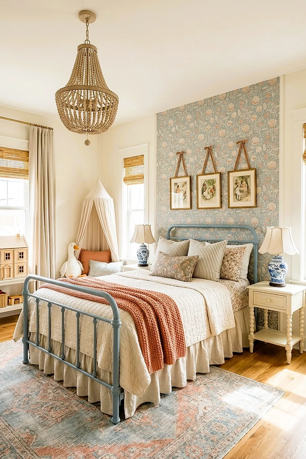 Girl's bedroom featuring a light blue metal bed with cream bedding and multiple patterned pillows, set against a blue floral wallpaper accent wall. The room includes beige curtains, woven shades on the windows, two cream-colored nightstands with blue and white ceramic lamps, a hanging white fabric canopy in the corner, hardwood floors with a vintage-style rug, and framed vintage art hung above the bed with leather straps.