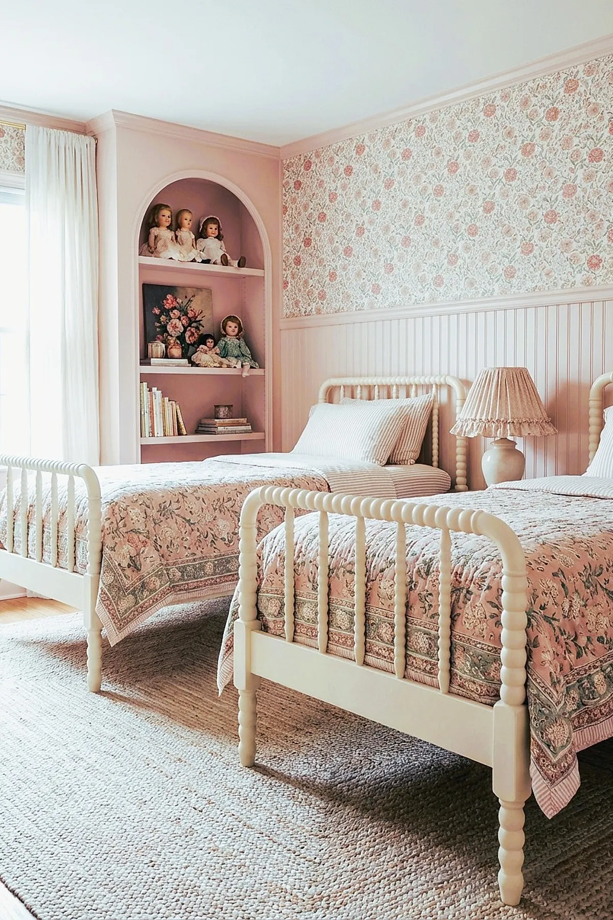 Twin girl bedroom with cream-colored spindle beds dressed in floral pink quilts, featuring beadboard wainscoting and floral wallpaper above, a built-in arched shelf painted pink holding vintage dolls, books, and decorative objects, complemented by a beige textured rug on wooden flooring.