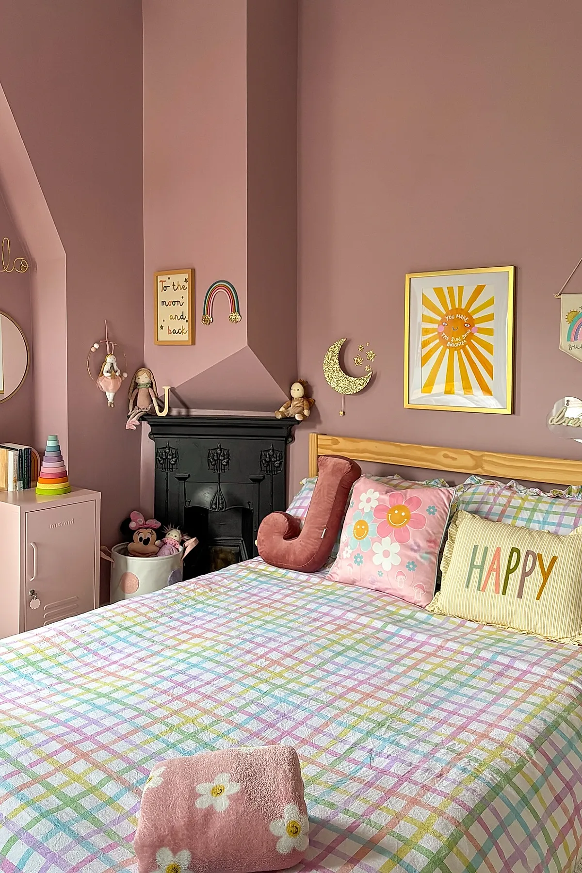 Big girl bedroom with pastel checkered bedding on a wooden bed frame, featuring decorative pillows including one with 'HAPPY' text, pink walls adorned with gold-framed sun artwork and glittery moon wall decor, alongside stuffed toys and colorful accessories.