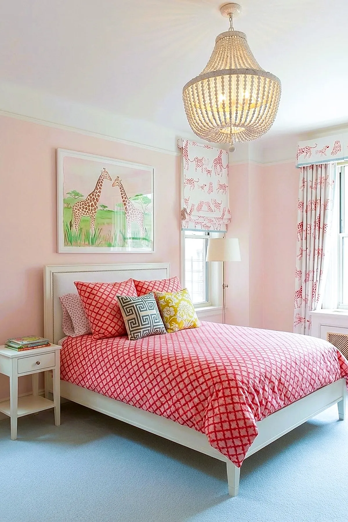 Girl's bedroom with soft pink walls, featuring a white bed dressed in a red diamond-patterned duvet and pillows, accented by a yellow floral pillow and a black and white geometric pillow, giraffe artwork above the bed, and matching curtains and roman shades with red animal prints on white background.