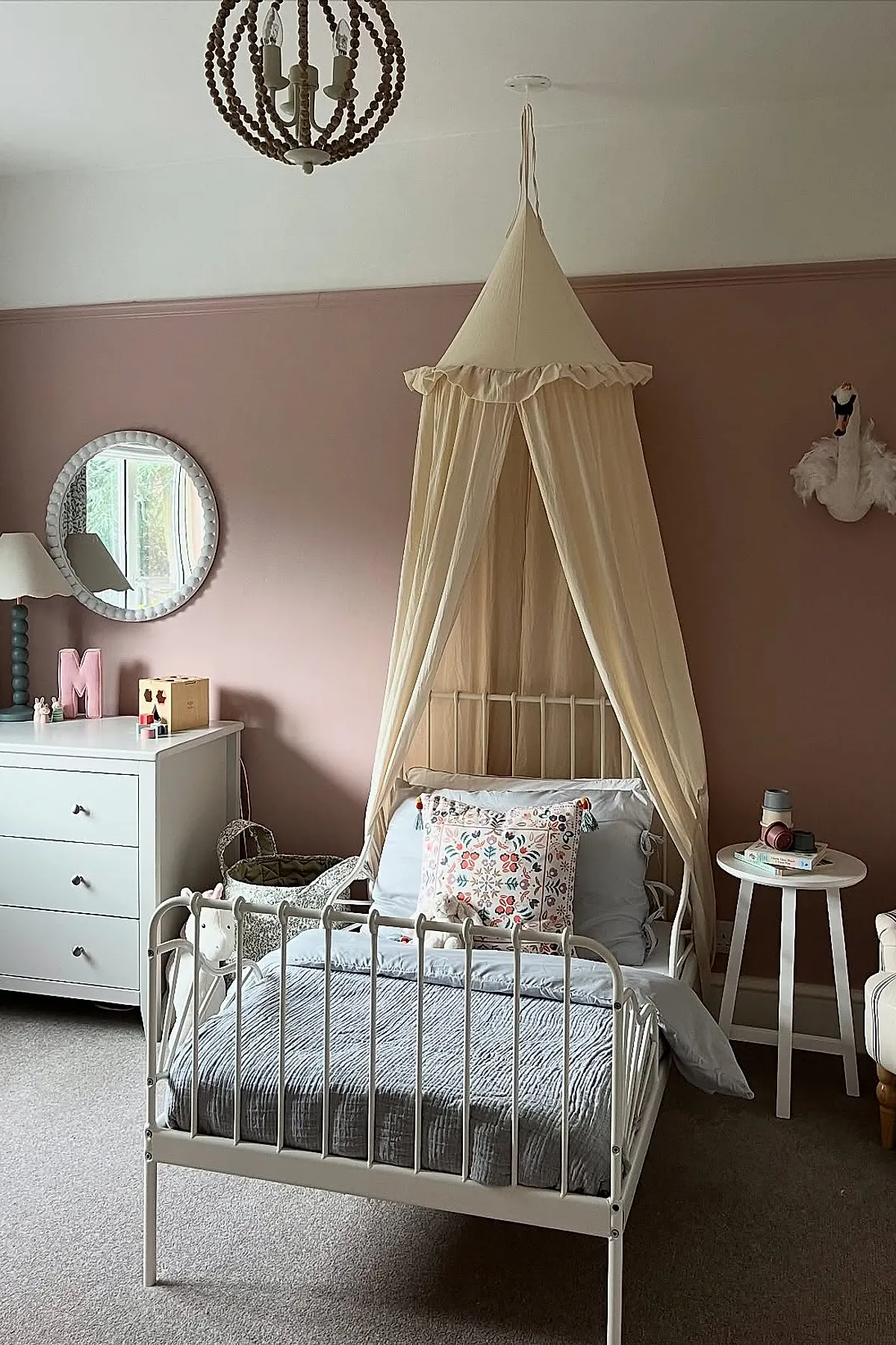 Girl's bedroom with pink walls featuring a white metal bed frame topped with an off-white fabric canopy, decorated with floral accent pillows and soft bedding, complemented by white dresser, round mirror, wooden beaded chandelier, and a swan wall decor.