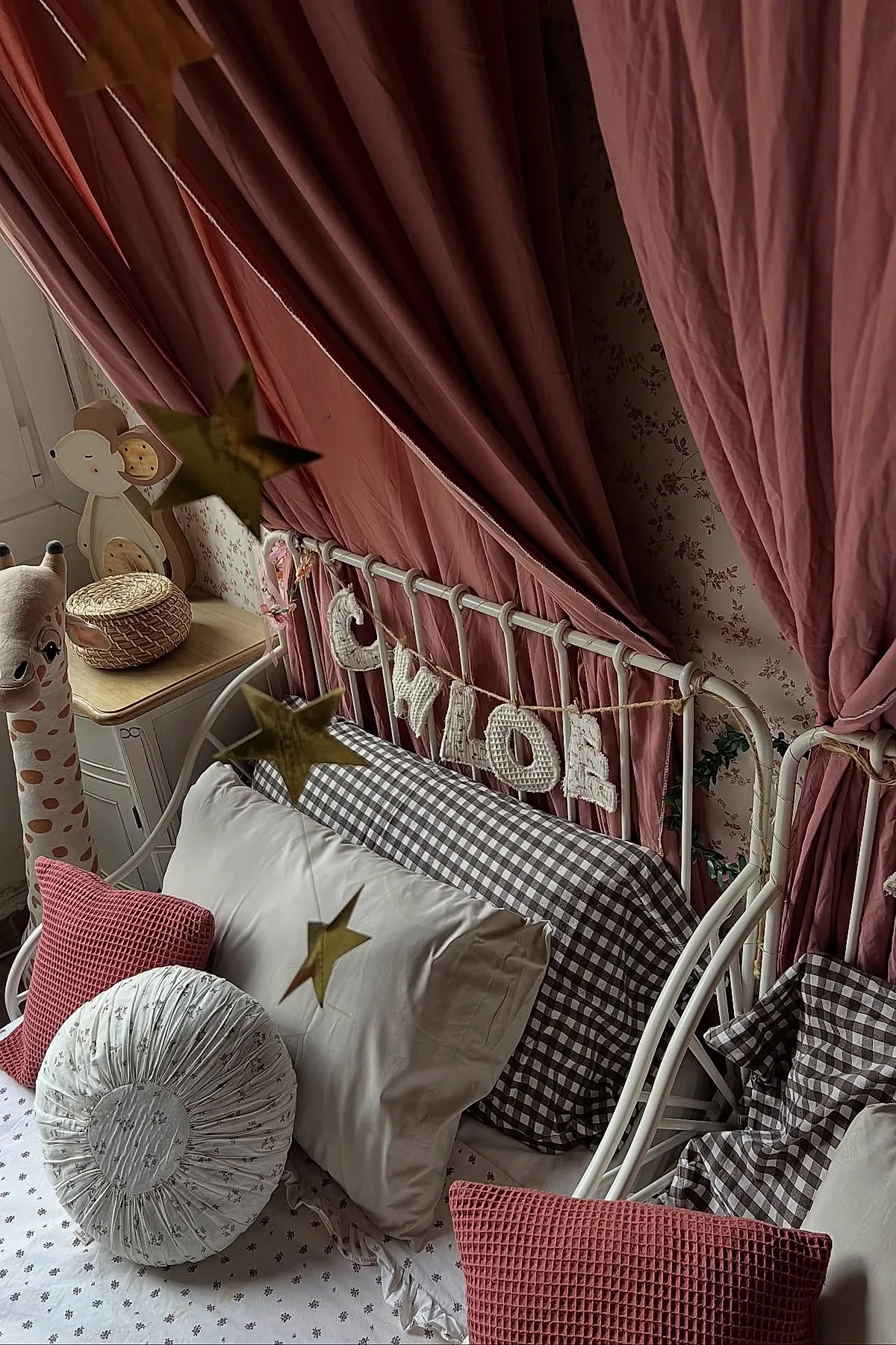Girl's bedroom featuring a white metal bed frame with gray and white gingham pillowcases, dusty rose curtains tied back on each side, textured pink square pillows, a round white cushion, star-shaped hanging decorations, floral wallpaper in soft tones, and wooden nightstand with woven basket.