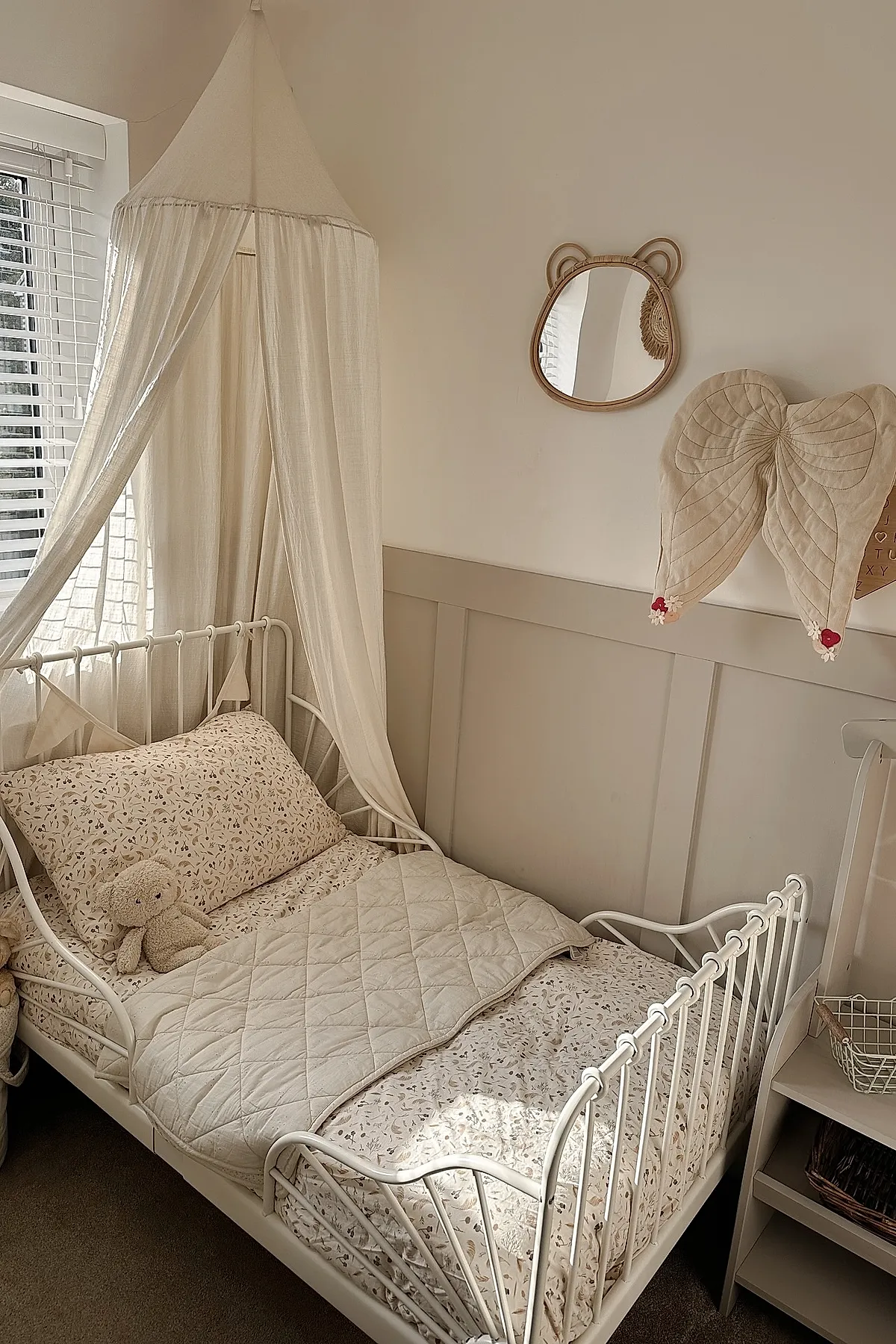 Girl's bedroom featuring a white metal bed with floral patterned bedding in neutral tones, topped with a sheer white fabric canopy, accompanied by beige paneled walls, a teddy bear on the bed, and cute wall decor shaped like wings and an animal face mirror.