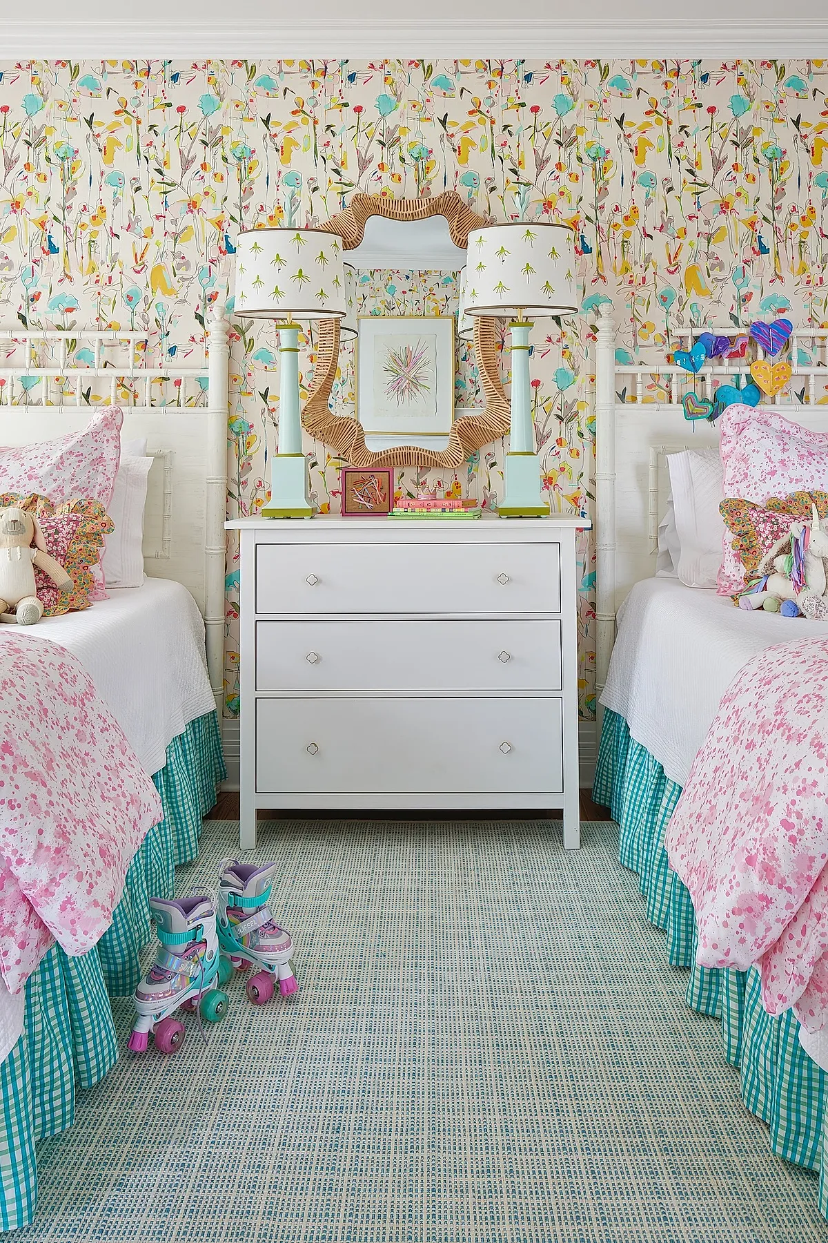 Twin girls' bedroom with white wooden beds dressed in pink floral bedding and green gingham bed skirts, colorful floral wallpaper covering the walls, a white three-drawer dresser topped with two matching lamps with patterned shades, a decorative mirror above, and pastel roller skates on a textured blue rug.