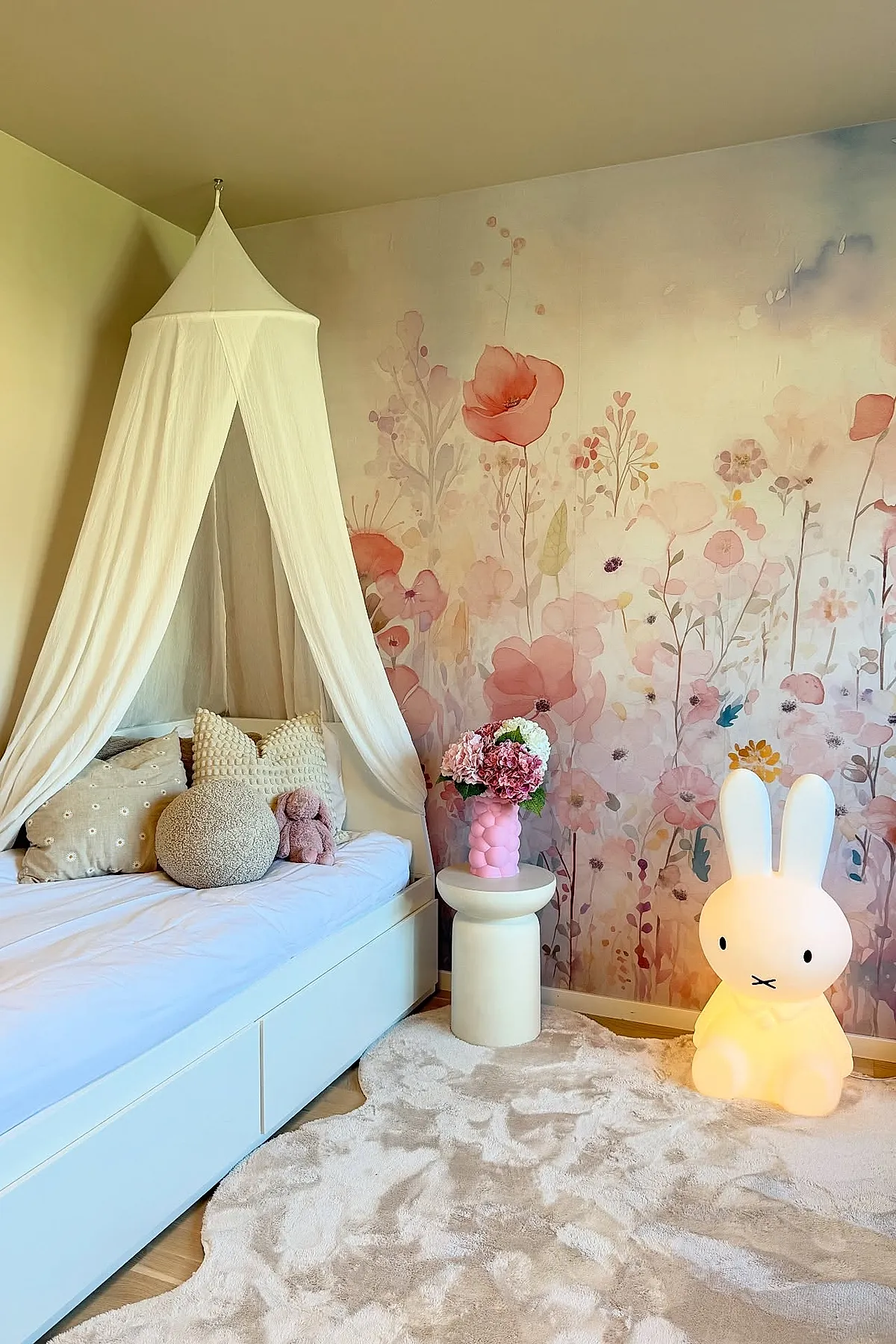 Girl's bedroom featuring a white canopy bed with soft beige and textured pillows, set against a wall covered in a pastel floral mural with pink and purple tones, accompanied by a small round white side table holding a pink vase of flowers, and a glowing bunny lamp on a plush cream-colored rug.