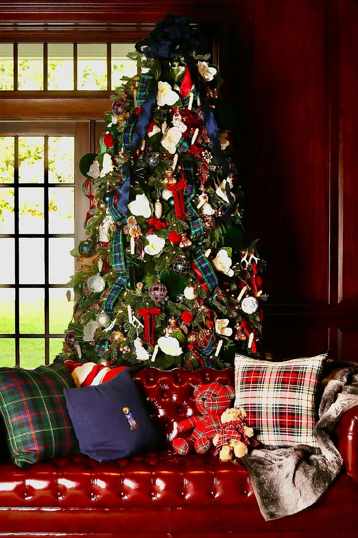 A traditional christmas tree decorated with blue and green tartan ribbon garlands, large white magnolia flowers, red berries, assorted ornaments, set behind a red leather sofa adorned with plaid pillows and matching teddy bears dressed in plaid.