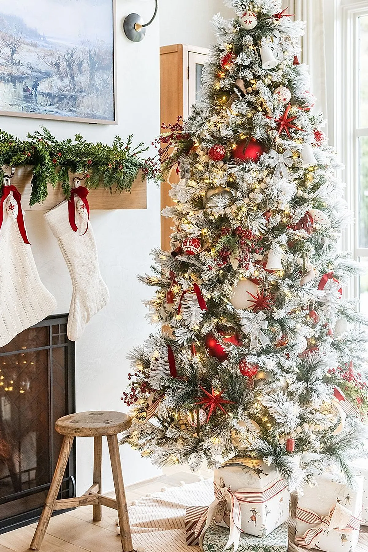 Traditional frosted Christmas tree decorated with red baubles, white bell ornaments, white bows, and red berry sprigs illuminated by warm white lights next to a fireplace mantel adorned with green garland and cream knitted stockings tied with red ribbons.