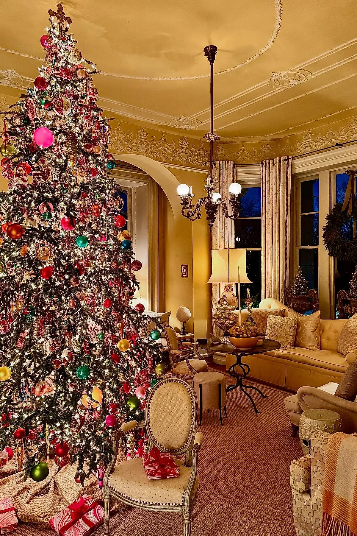 traditional christmas tree densely decorated with multicolored glass ornaments including red, green, gold, and pink baubles illuminated by warm white string lights in a warmly lit living room featuring ornate ceiling details, cream curtains, and classic upholstered furniture.