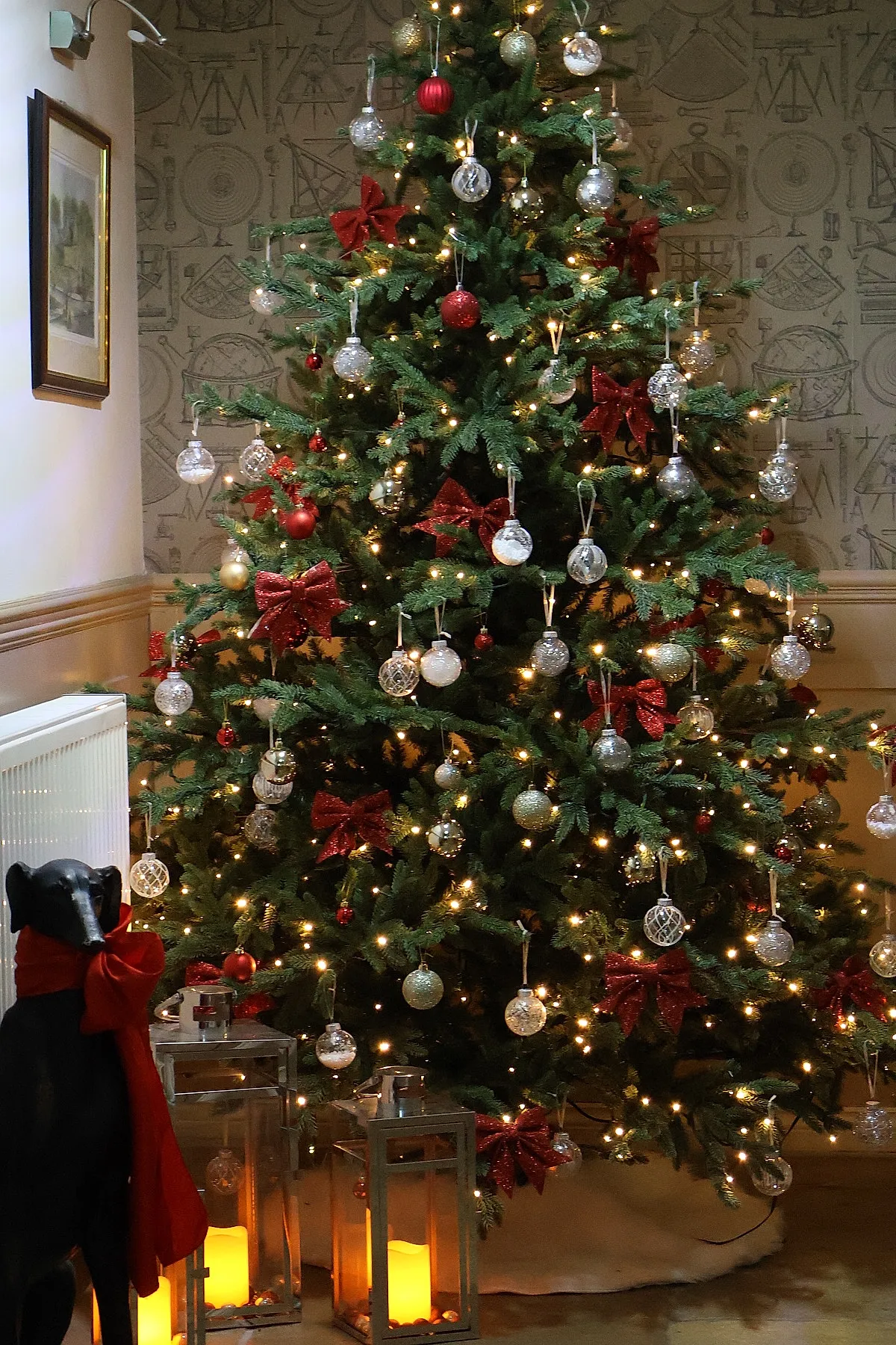 A traditional christmas tree decorated with red glitter bows, clear and gold glass ball ornaments, warm white string lights, and nearby metal lanterns holding glowing candles.