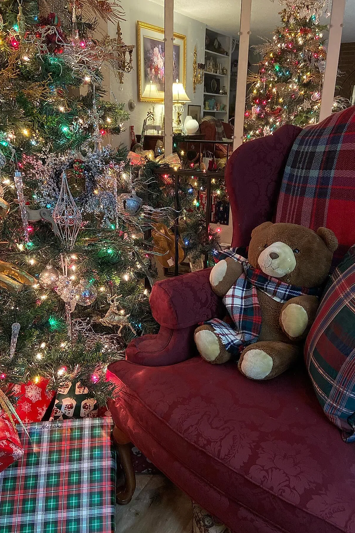 traditional christmas tree decorated with multicolored string lights, silver and glass ornaments, and red tartan ribbons, reflected in mirrors behind a red patterned wingback chair holding a brown teddy bear wearing a matching red tartan scarf.