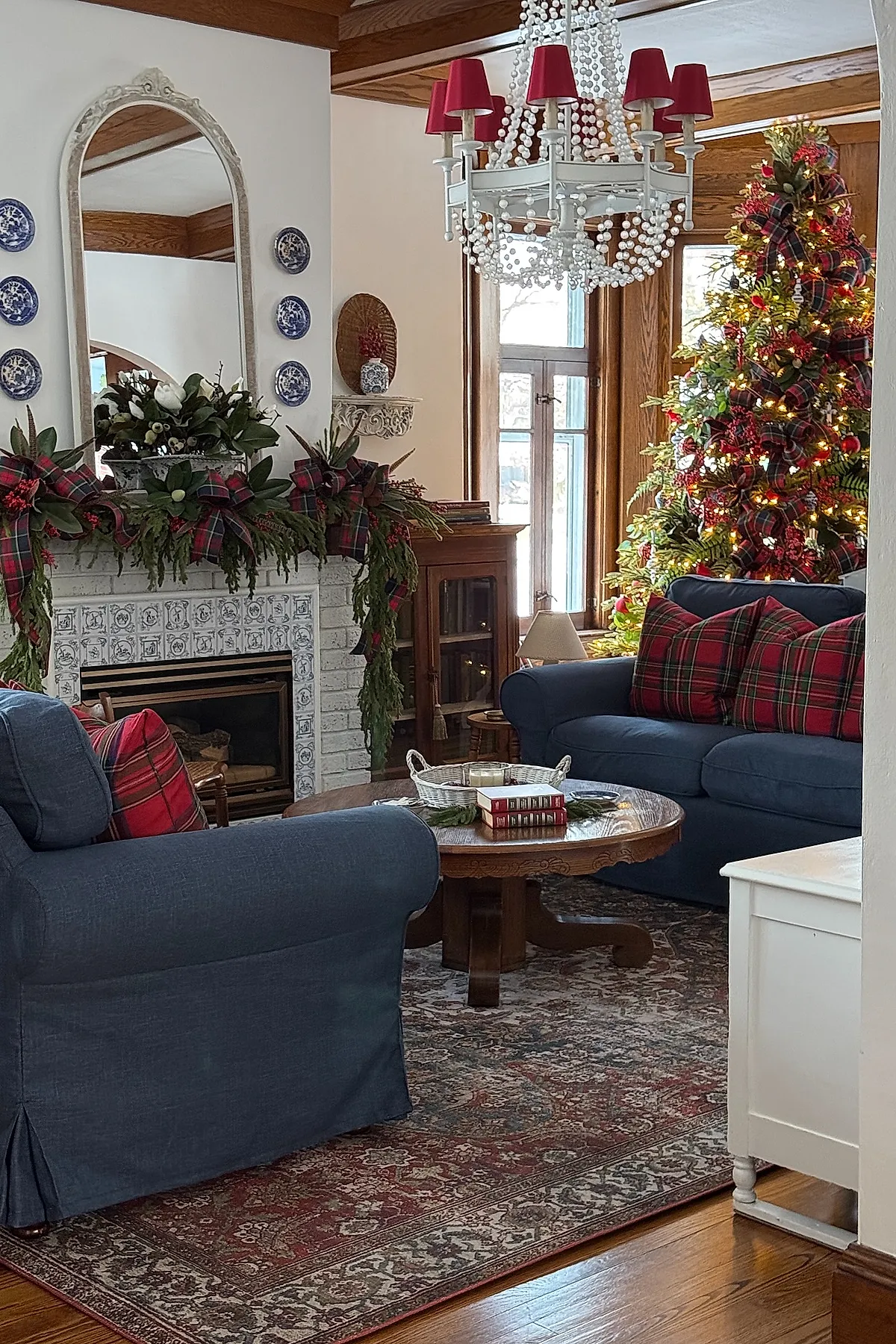 traditional christmas tree decorated with red and green tartan ribbon garlands, clusters of red berry picks, warm white string lights, standing near blue sofas accented with matching tartan pillows in a cozy living room.