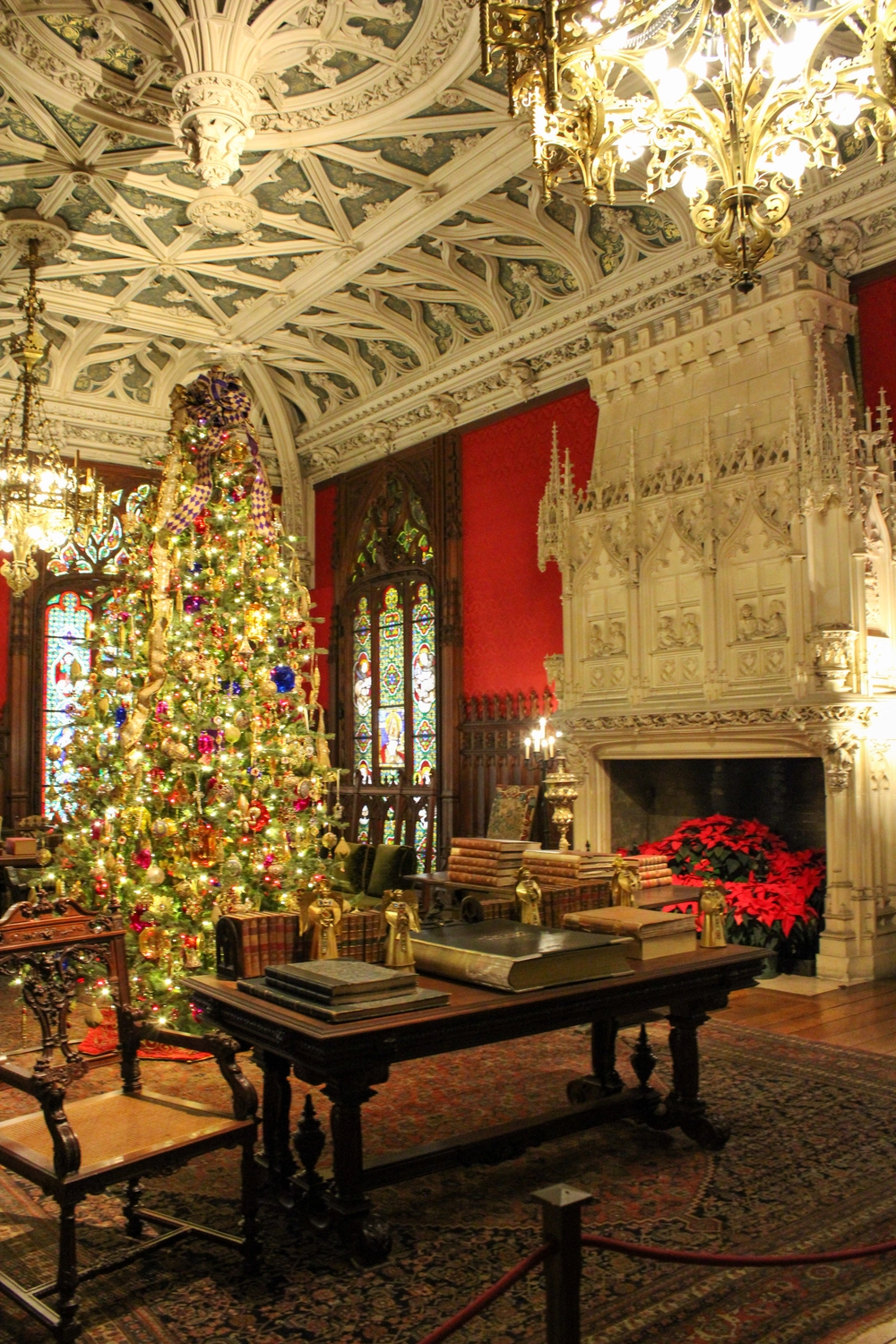 traditional Christmas tree decorated with gold ribbon, red and green ornaments, and warm lights in an ornate historic room with stained glass windows and red walls