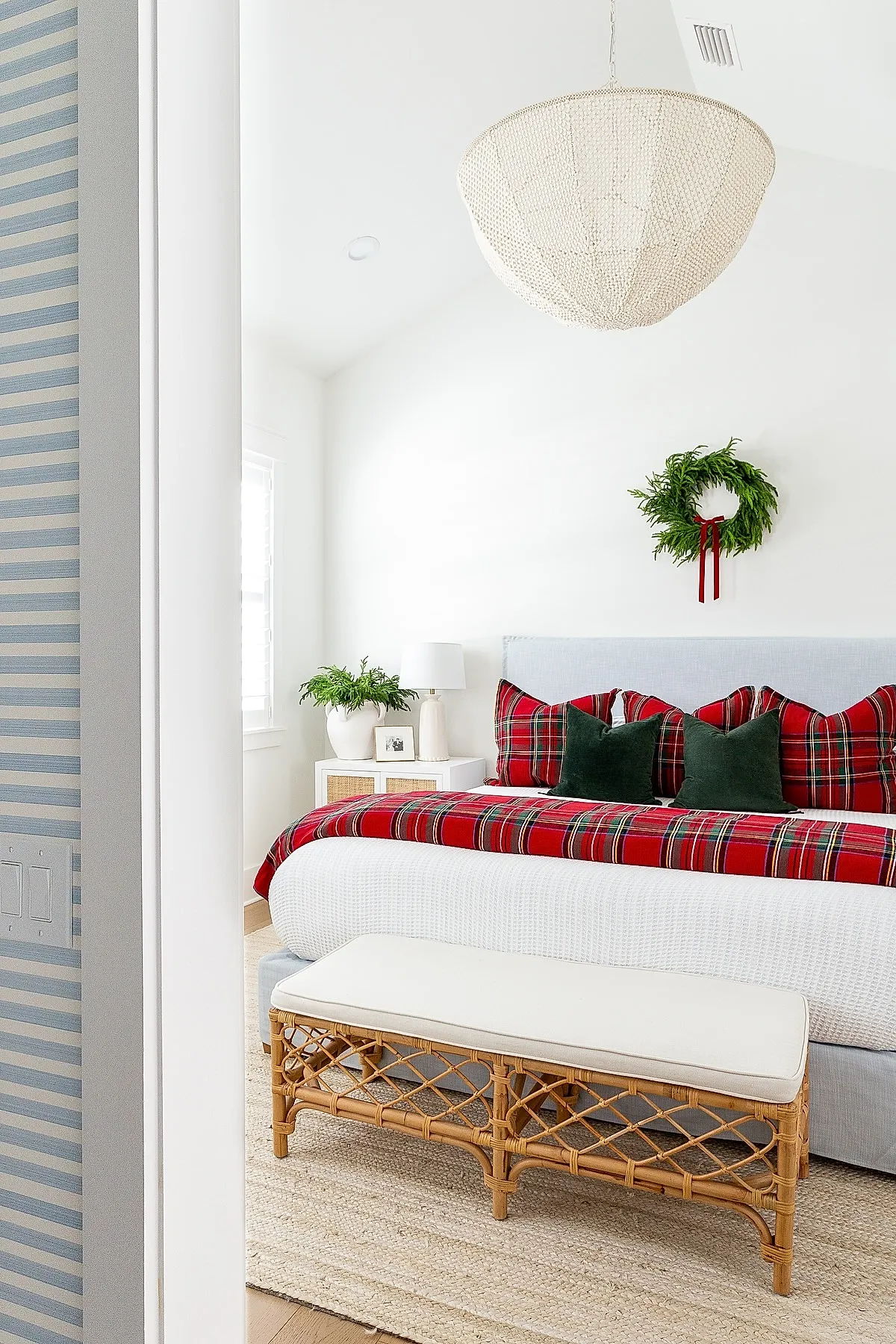 Bright white bedroom featuring a light blue upholstered bed with red plaid pillows and throw blanket, green velvet accent pillows, a green wreath with red ribbon hanging above the headboard, a white nightstand with lamp and potted plant, wicker bench with cream cushion at foot of bed, natural woven rug on wooden floor.