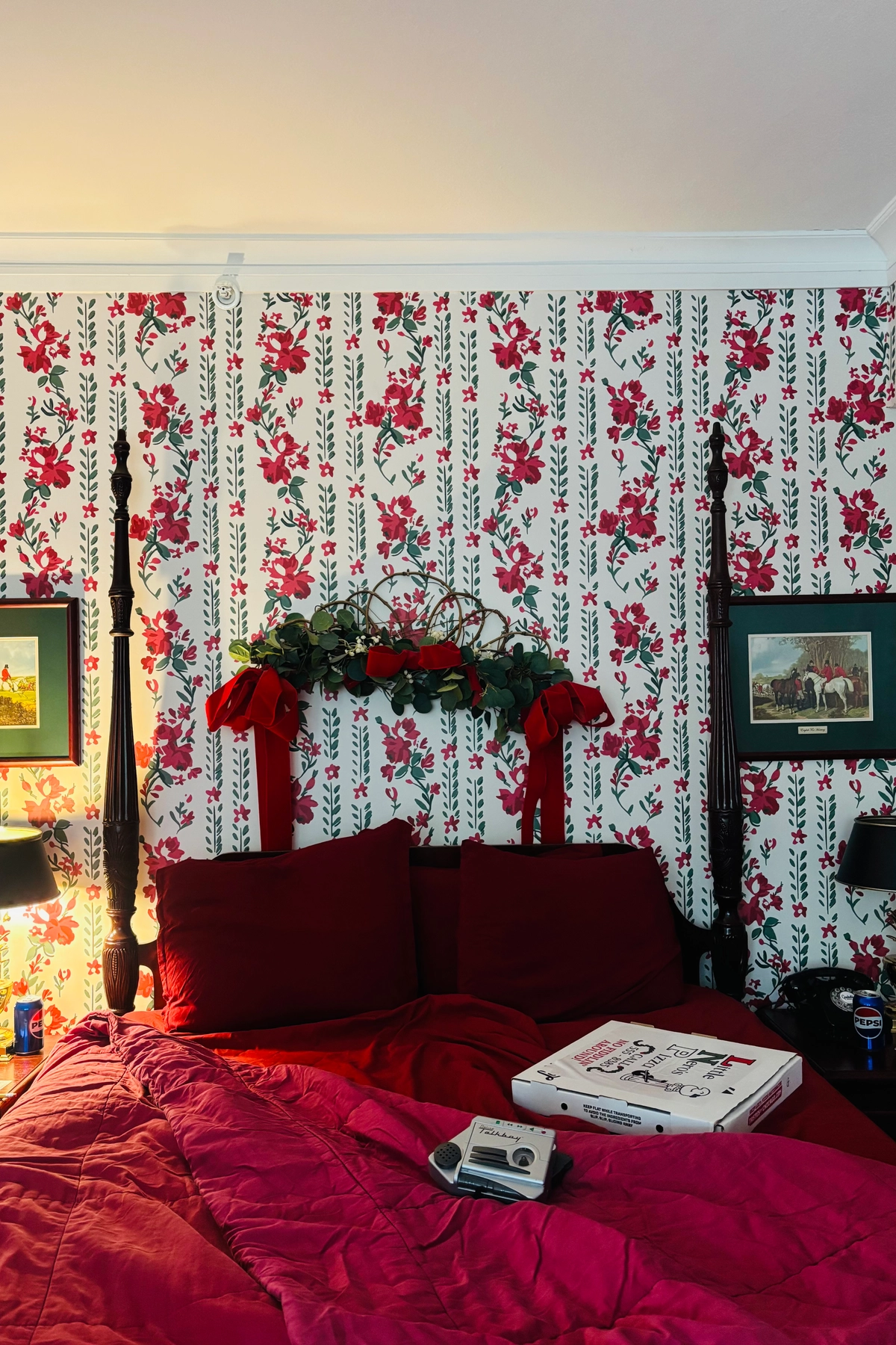 A Christmas bedroom decorated with bold red bedding, a floral red and green wallpaper, dark wood four poster bed, and a festive greenery swag with red velvet ribbons above the headboard.