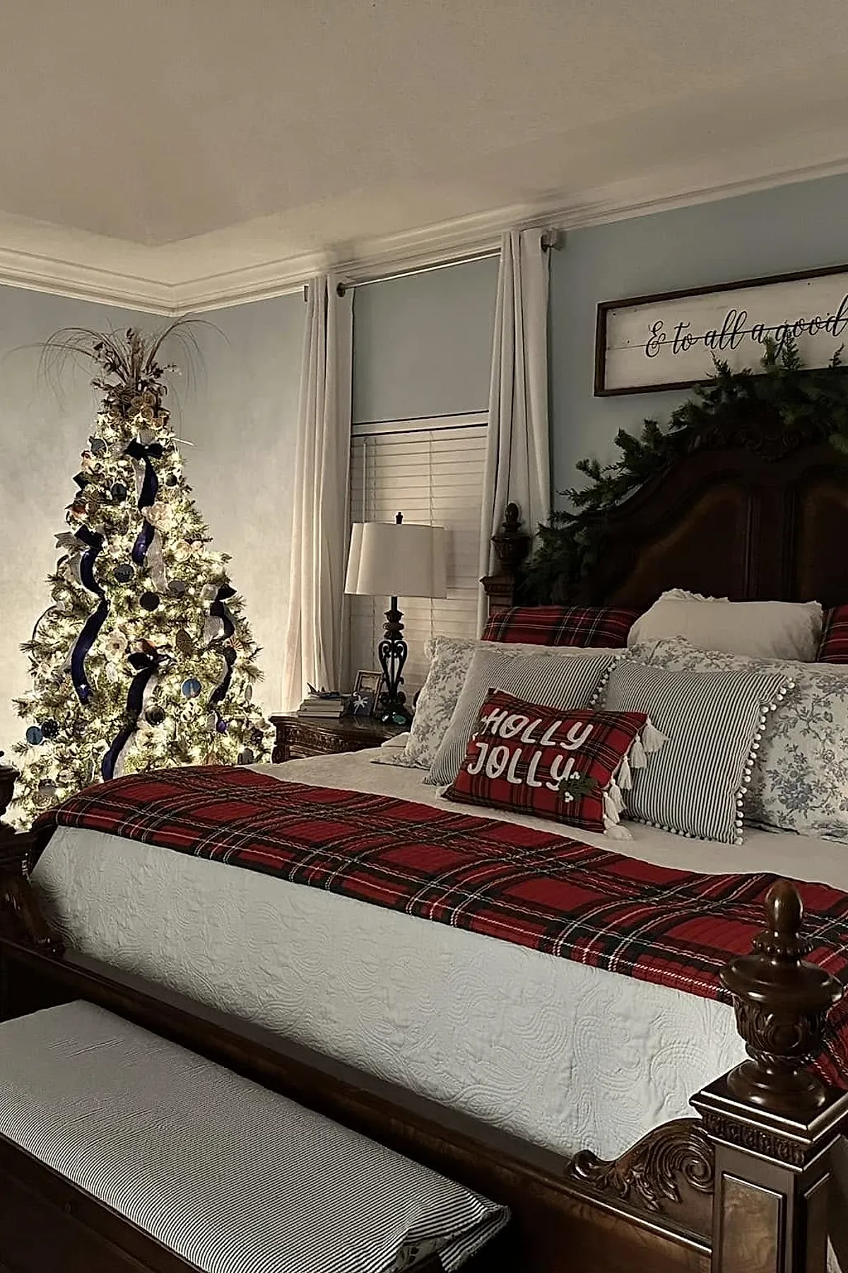 Christmas bedroom featuring a dark wooden bed with an ornate headboard decorated with greenery, bedding layered with red plaid blankets and pillows including one that says 'Holly Jolly', a warmly lit white Christmas tree adorned with blue ribbon, cream curtains framing the window, and soft ambient lighting.