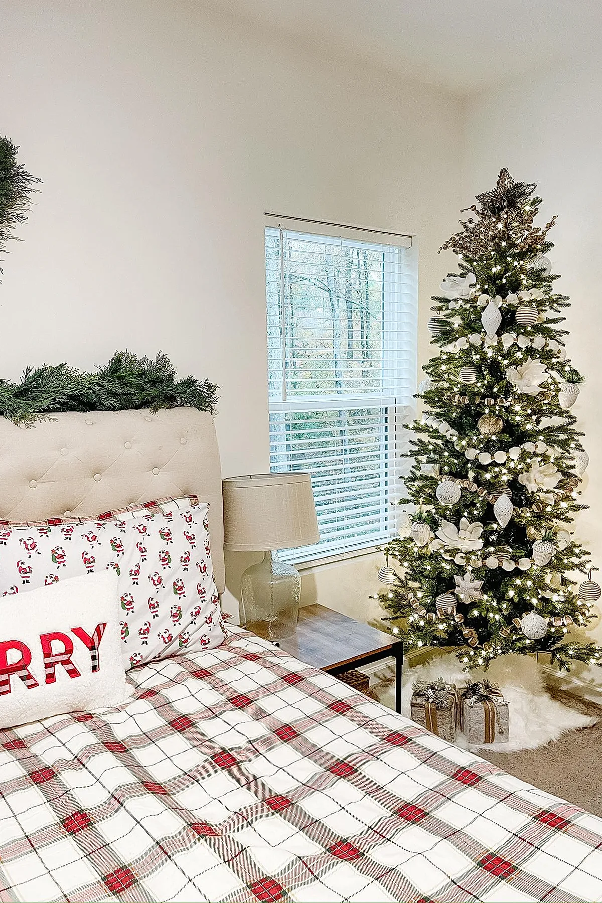 Christmas bedroom with a beige tufted headboard decorated with greenery, red and white plaid bedding, pillows featuring Santa Claus prints and a white pillow with 'MERRY' in red letters, a white Christmas tree adorned with gold ornaments and lights, a wooden side table with a clear lamp, all set against white walls and window blinds.
