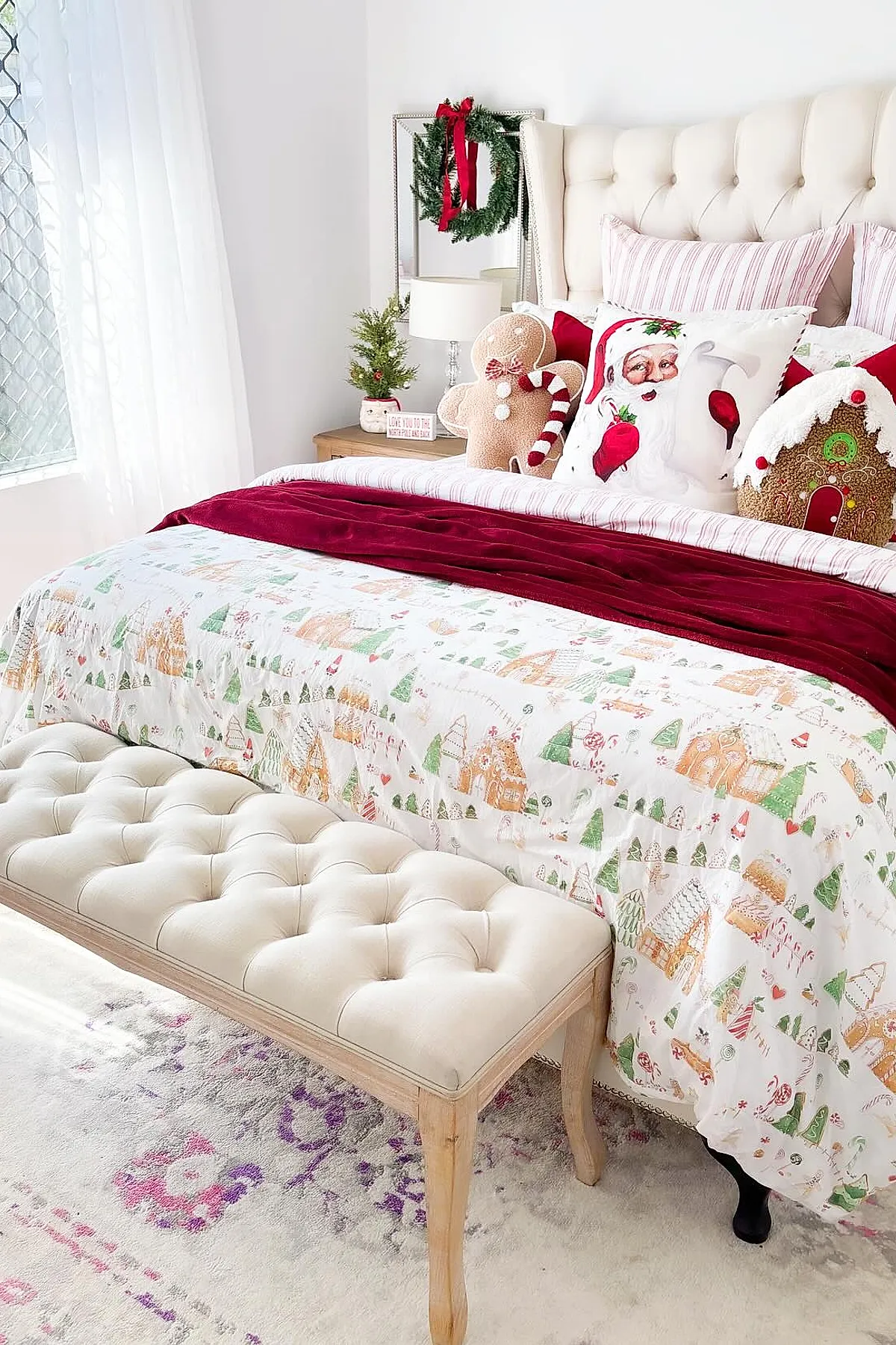 Christmas bedroom with tufted cream headboard, festive bedding featuring pastel holiday village prints, red velvet throw blanket, pillows shaped like Santa Claus, gingerbread man, and gingerbread house, small decorated pine tree on wooden nightstand with lamp and mirror behind adorned with green wreath tied with red ribbon.