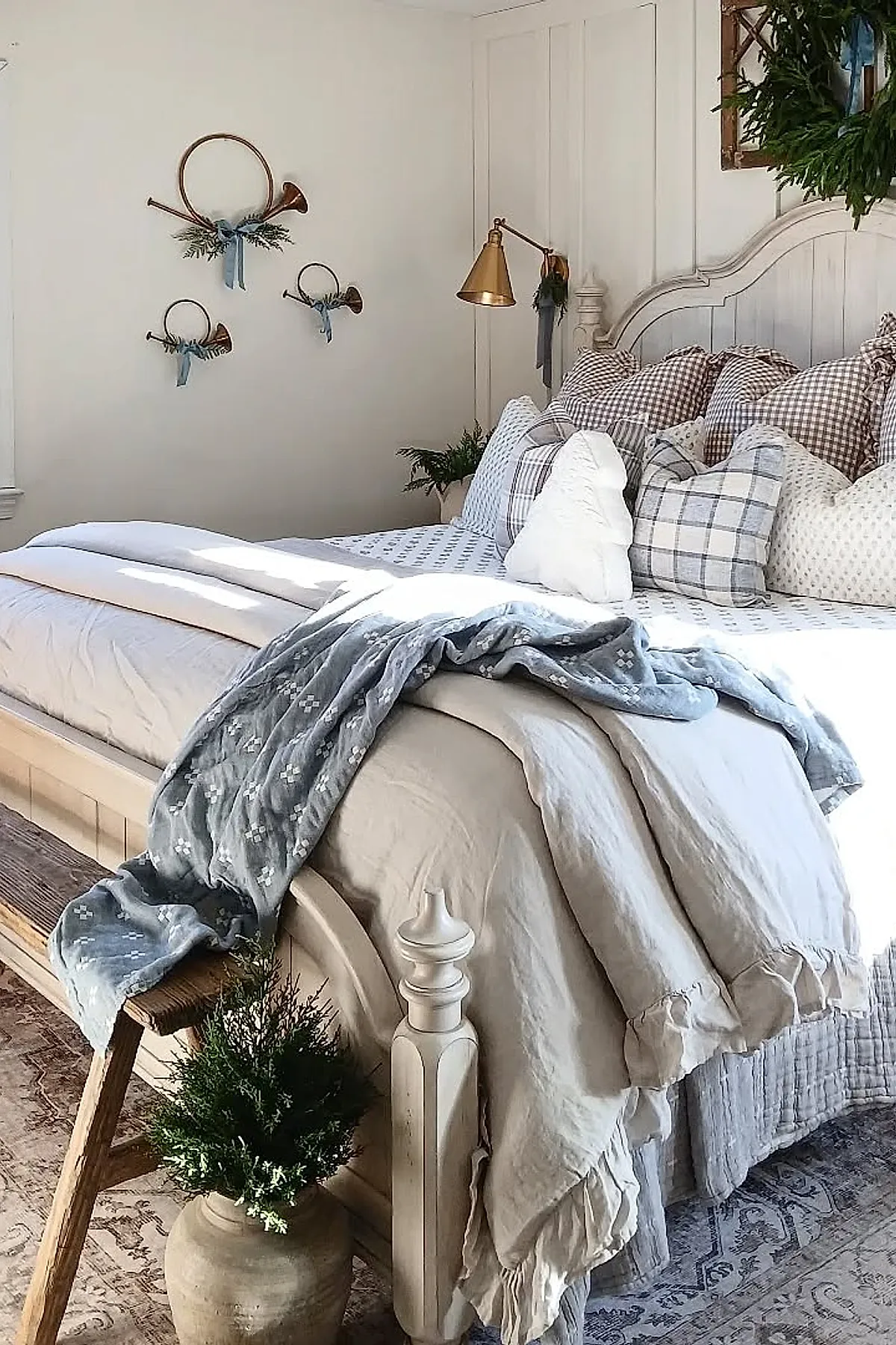 Christmas bedroom featuring a white wooden bed with beige and brown gingham pillows, cream bedding, a light blue patterned throw draped over the footboard, vintage brass horns decorated with greenery and blue ribbons mounted on a white wall, a wooden bench at the foot of the bed, and a green wreath hanging above the headboard.
