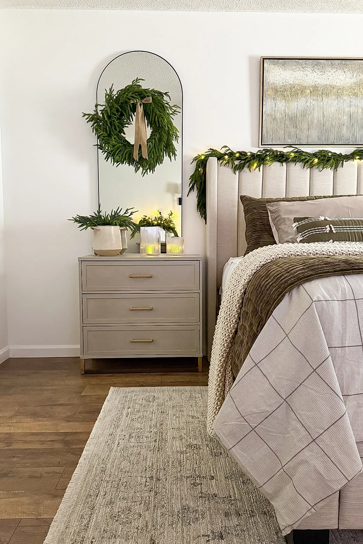 Neutral-toned Christmas bedroom with a beige upholstered bed, green garland draped over the headboard with string lights woven in, a tall arched mirror decorated with a fresh green wreath hanging from a beige ribbon, light wood three-drawer dresser topped with a beige ceramic pot holding greenery and two lit candles in glass holders.