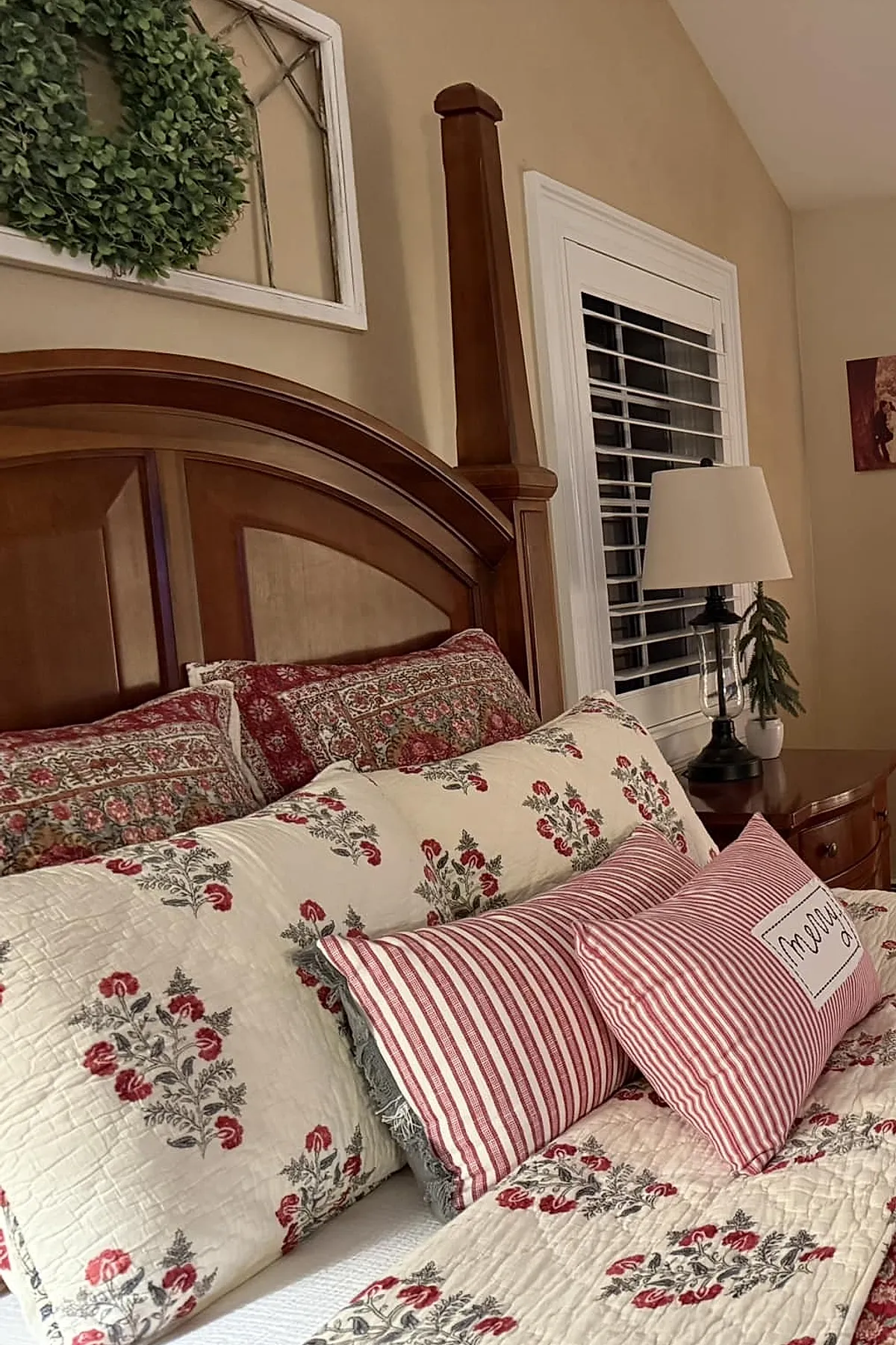 Christmas bedroom featuring a wooden headboard with beige walls, bedding with red floral patterns, matching floral pillow shams, two red and white striped accent pillows including one with the word 'merry', a lamp on a wooden nightstand, and a green wreath hanging above the bed.
