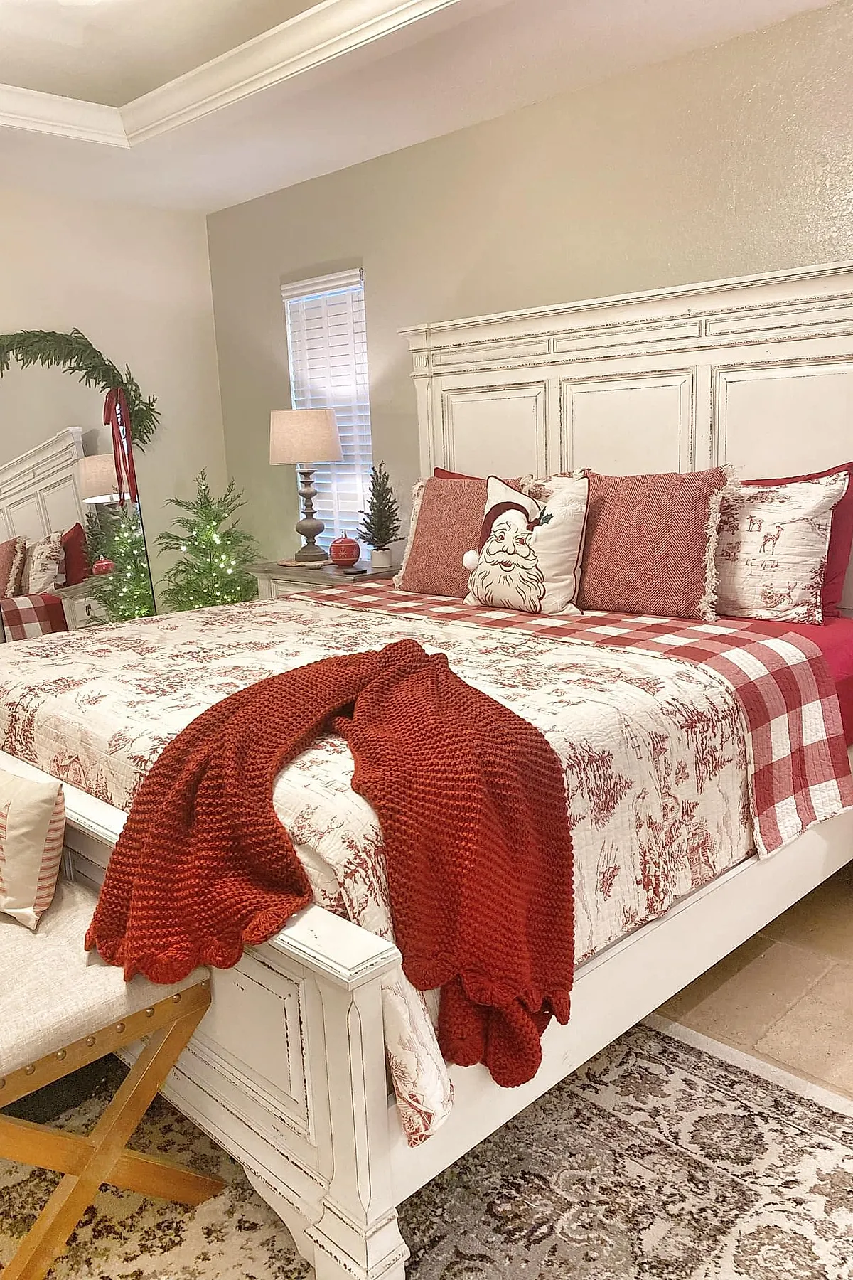 Christmas bedroom featuring a white wooden bed with distressed finish, red and white plaid bedding layered with toile patterned quilt, three textured red pillows, one pillow with a vintage Santa face print, and a chunky knit red blanket draped over the footboard. Nearby is a lamp on a bedside table with small greenery accents.