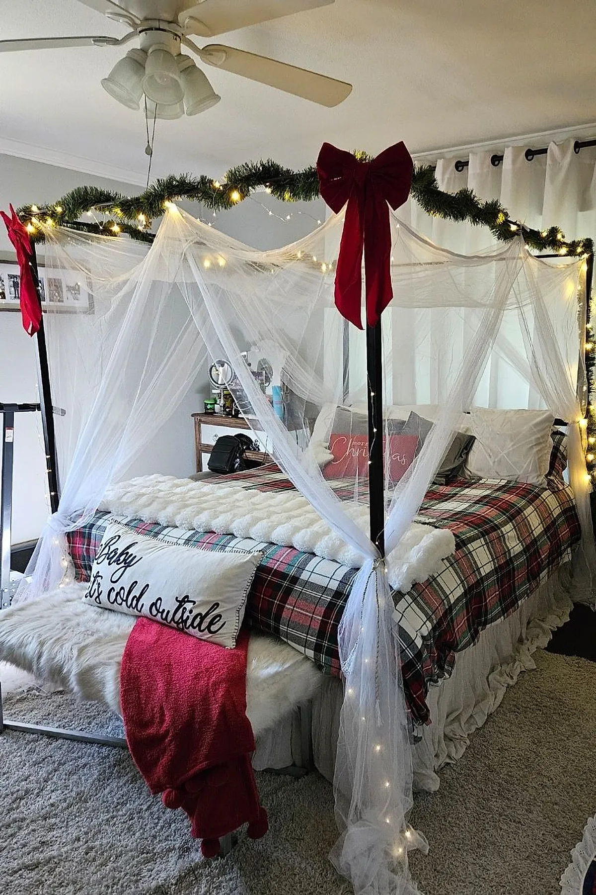 Christmas bedroom with a four-poster bed decorated with white sheer canopy drapes tied at the posts, green garland wrapped around the top frame with small warm string lights, large red bows on two corners, plaid bedding in red, white and green tones, a fluffy white throw blanket, pillows including one with 'Baby it's cold outside' text, and a furry bench at the foot of the bed topped with a red blanket.
