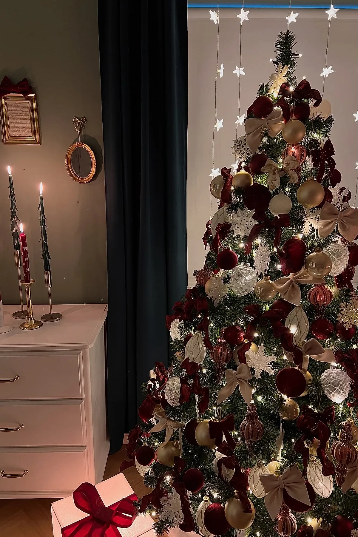 Christmas bedroom featuring a richly decorated Christmas tree with velvet burgundy bows, gold and white ornaments, snowflake decorations, and warm white string lights next to a white dresser holding tall green candles in brass holders, dark curtains, and star-shaped fairy lights hanging in the background.